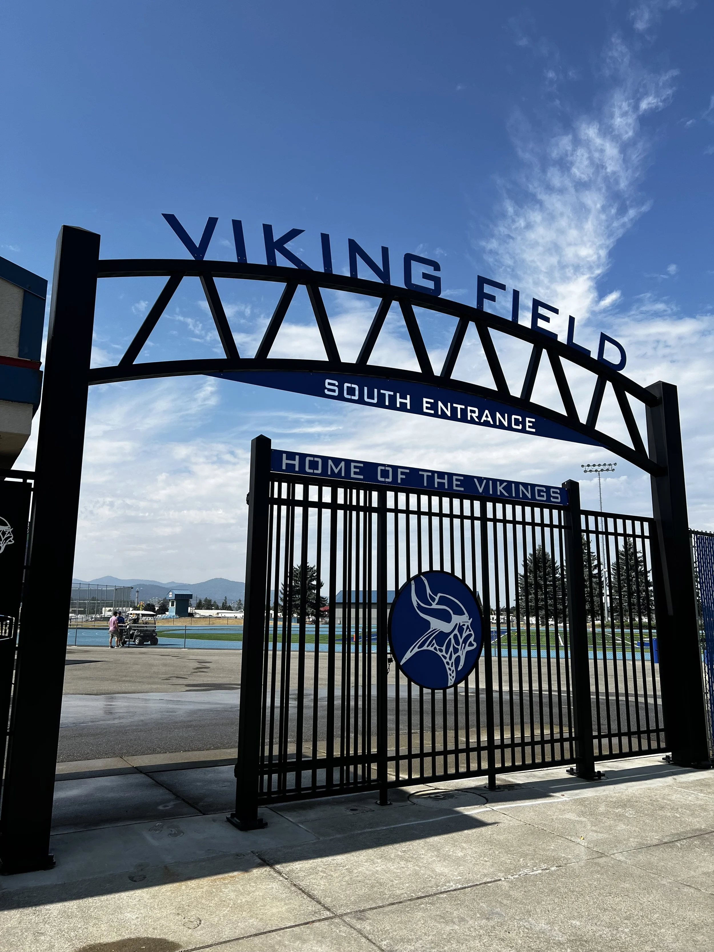 Entrance gate to Viking Field with signs reading 'VIKING FIELD', 'SOUTH ENTRANCE', and 'HOME OF THE VIKINGS' featuring a Viking helmet logo, at a sports facility on a partly cloudy day.