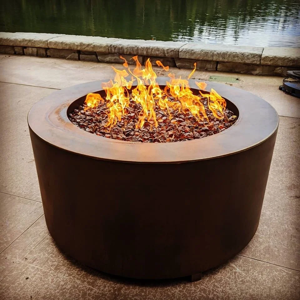 A round fire pit with flames and stones on top, situated near a body of water and paved surface.