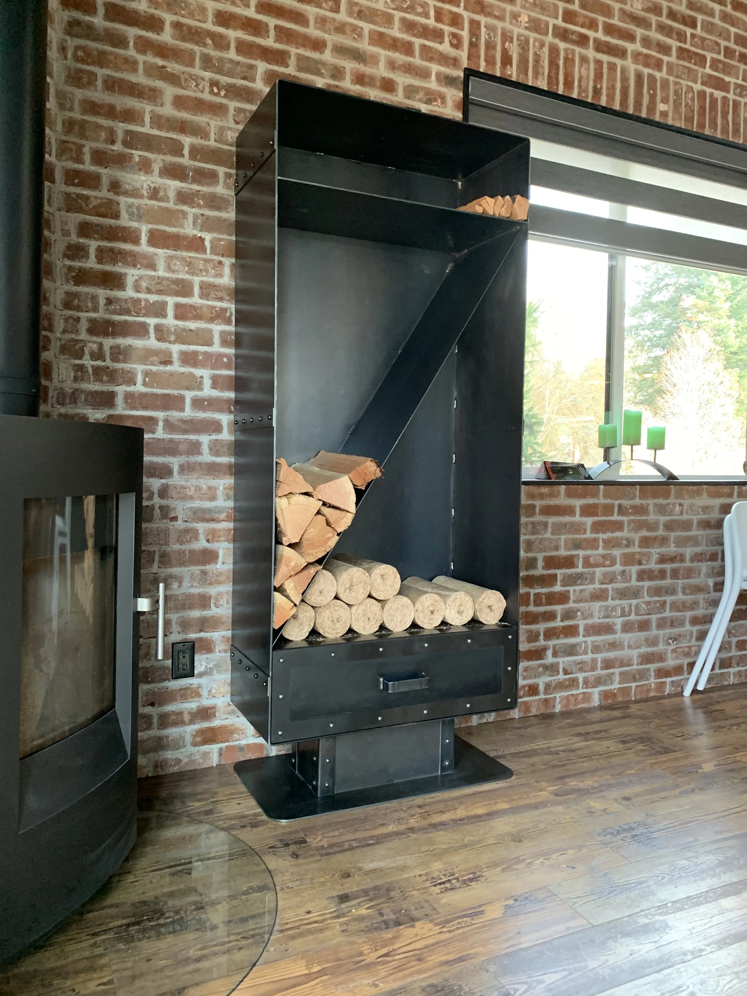A black metal wood storage cabinet filled with chopped and round firewood, positioned against a brick wall near a window.
