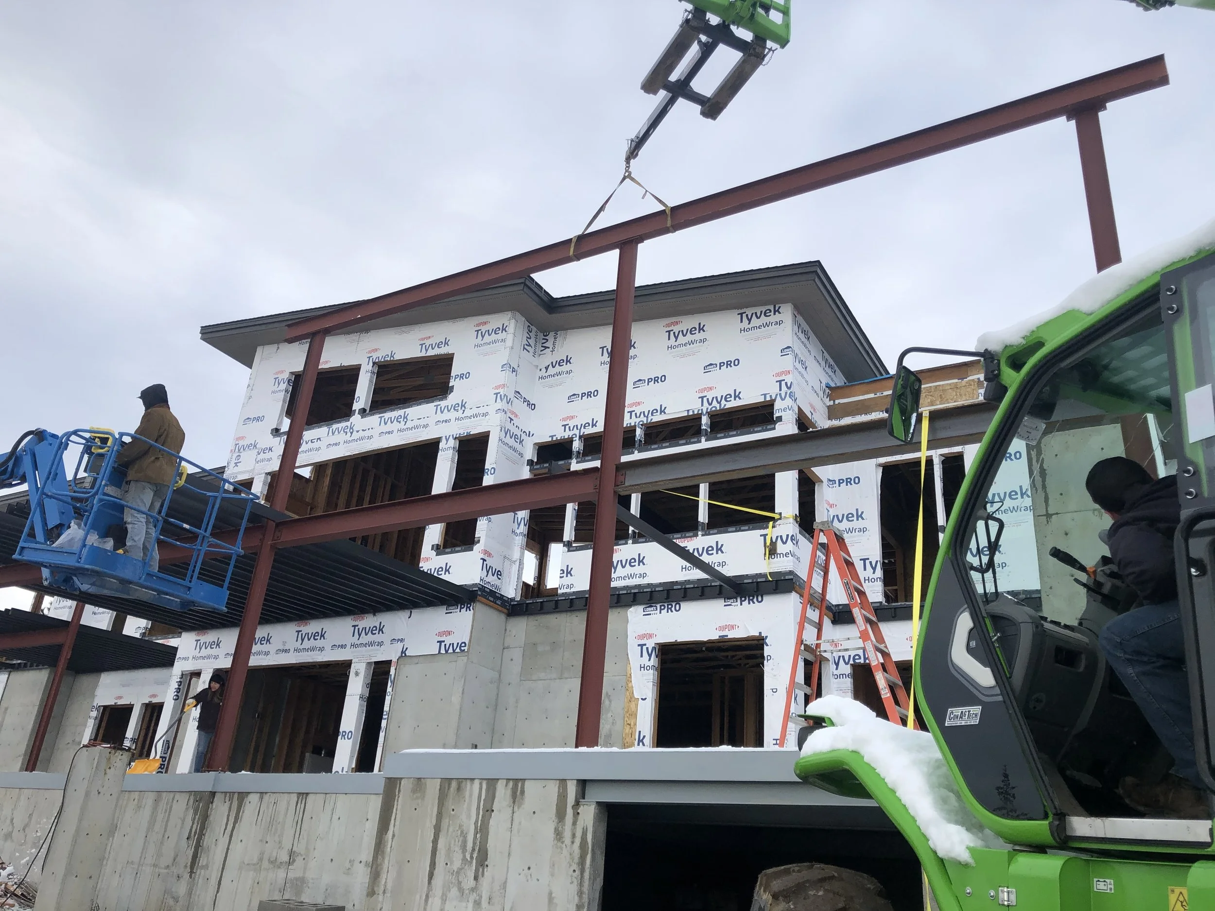 Construction workers building a multi-story house, with some on the ground and one on a lift, and steel beams being installed for the structure, with snow visible on parts of the machinery.