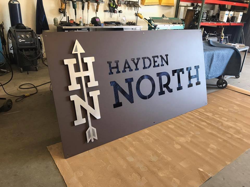 A custom metal sign on a work table with the words "HAYDEN NORTH" and an arrow design combining the initials H and N, in a workshop setting with tools and equipment in the background.
