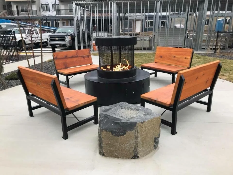 Four wooden benches with black metal frames arranged in a circle around a fire pit with a black metal mesh cover. A large rocks is in front of the benches.