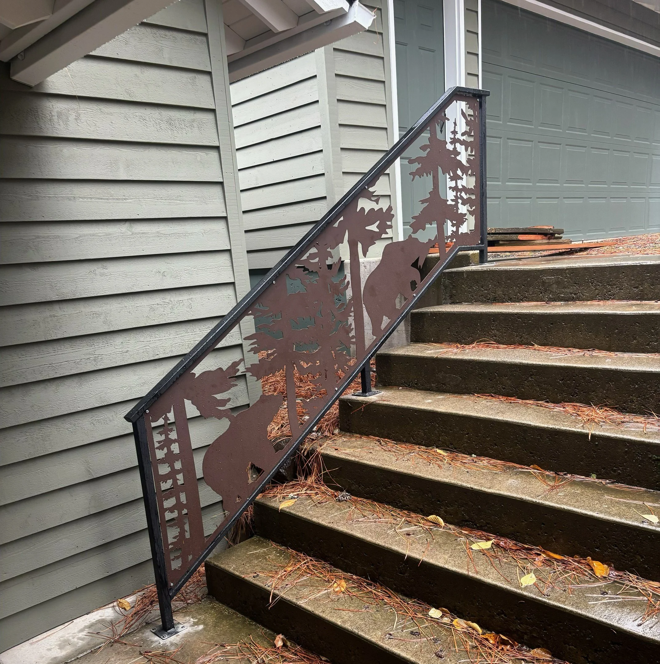 Exterior stairs with a decorative metal railing featuring cutout forest and bear designs, next to a building with light gray horizontal siding and a gray garage door, with fallen leaves and pine needles on the steps.