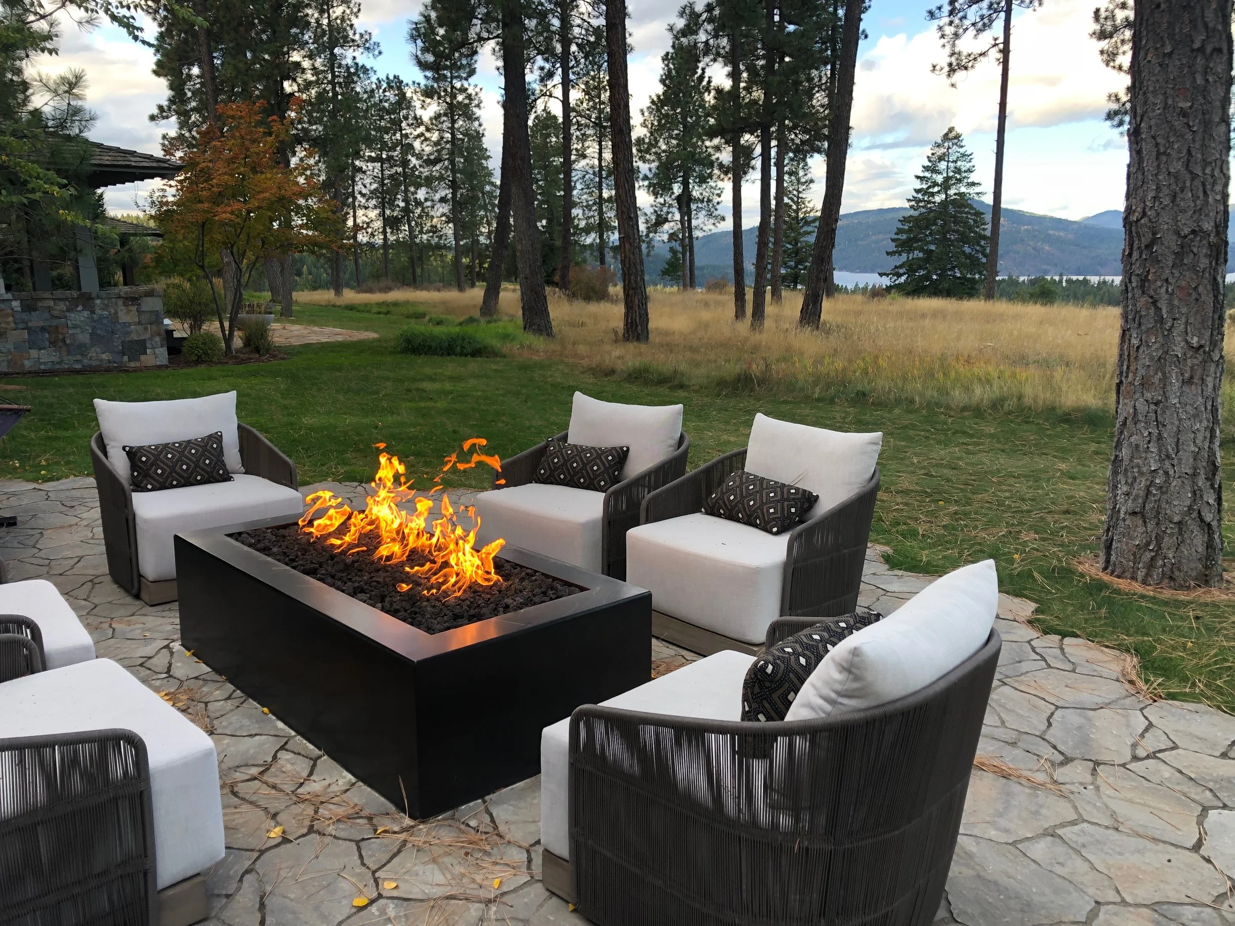 Outdoor patio with a firepit surrounded by black and white chairs, overlooking a grassy yard with trees and mountains in the background.