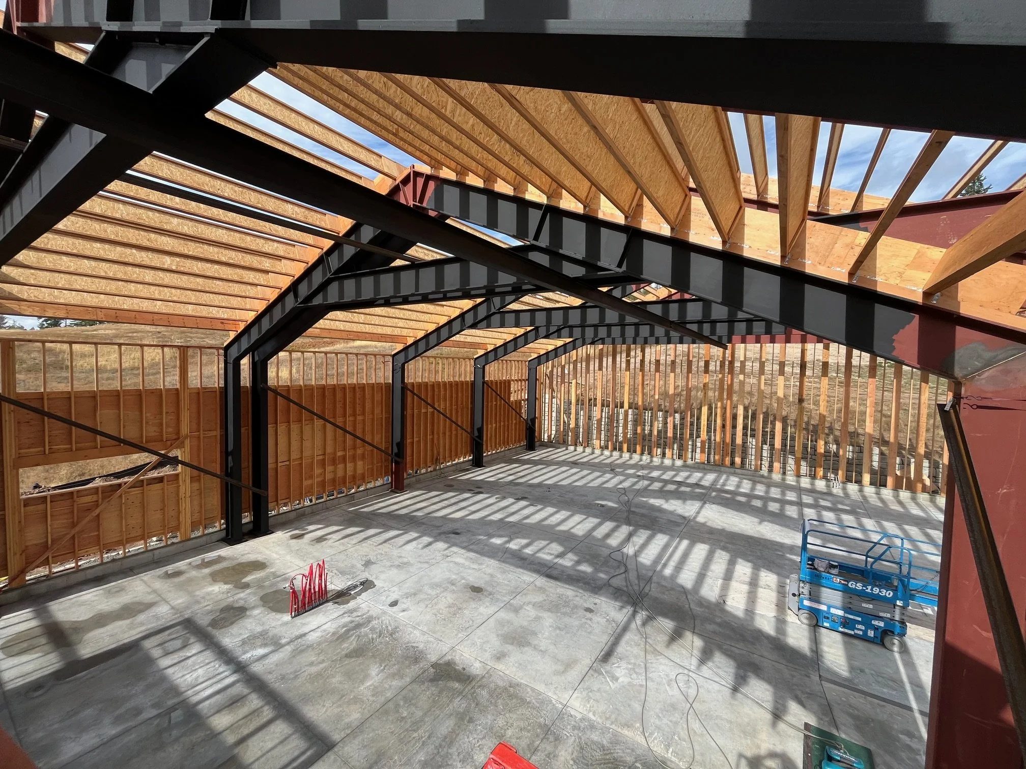 Construction site of a building with framing, steel beams, and partly installed roof panels, with construction tools and equipment on a concrete floor.