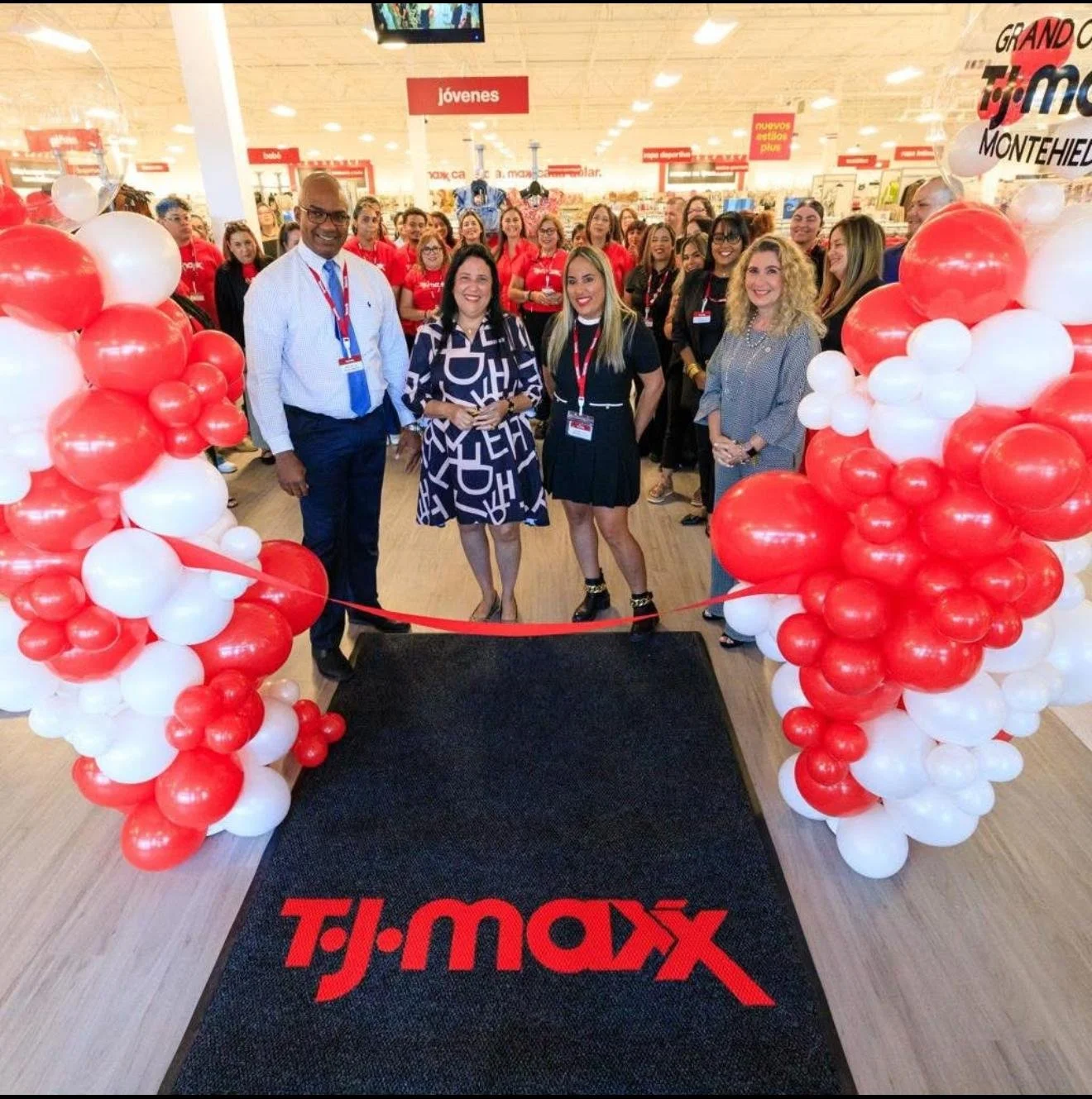 Group of people at the grand opening of a TJ Maxx store, standing behind a black mat with the store's logo, decorated with red and white balloons on either side.