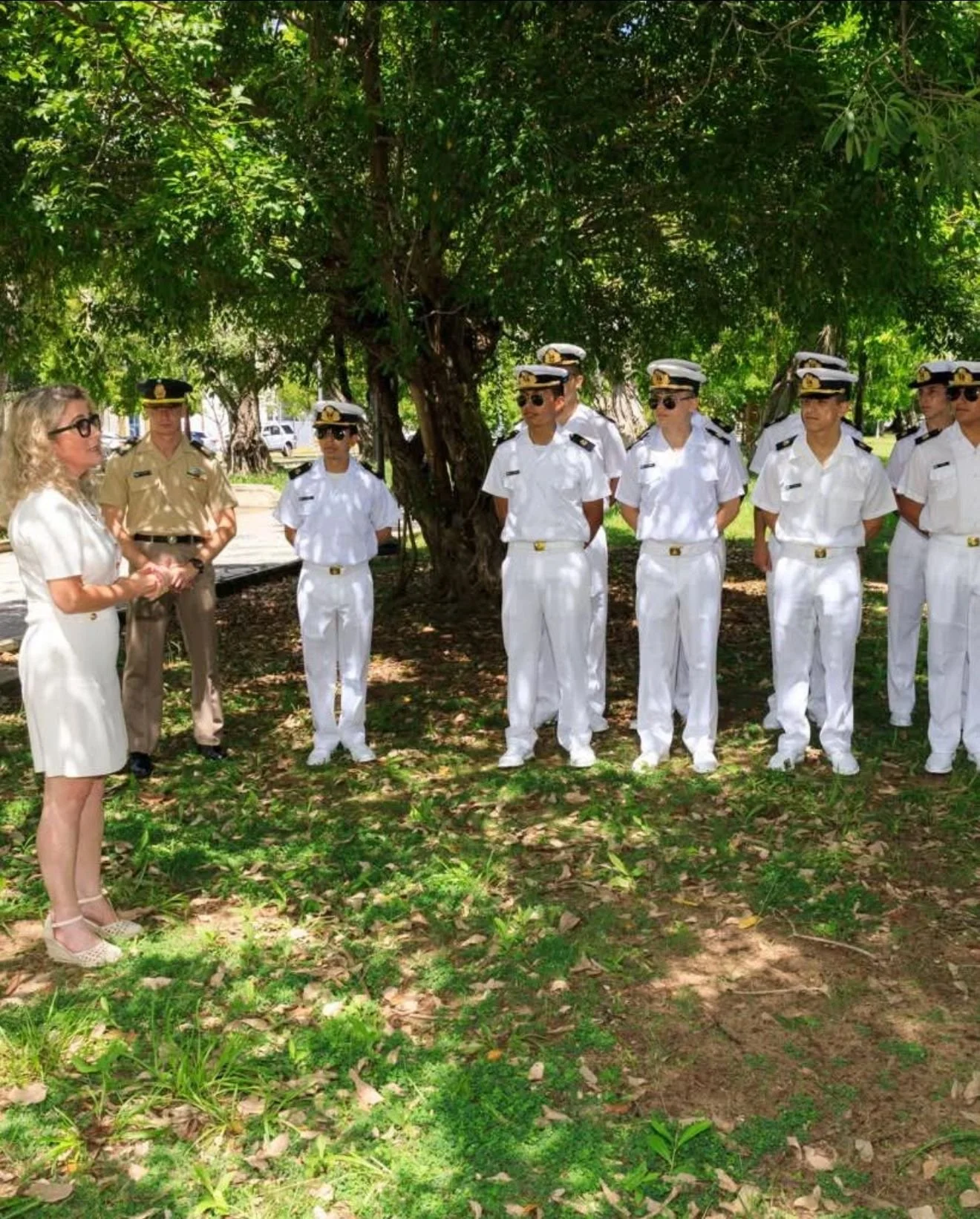 A woman speaking to a group of uniformed sailors outdoors beneath a large tree.
