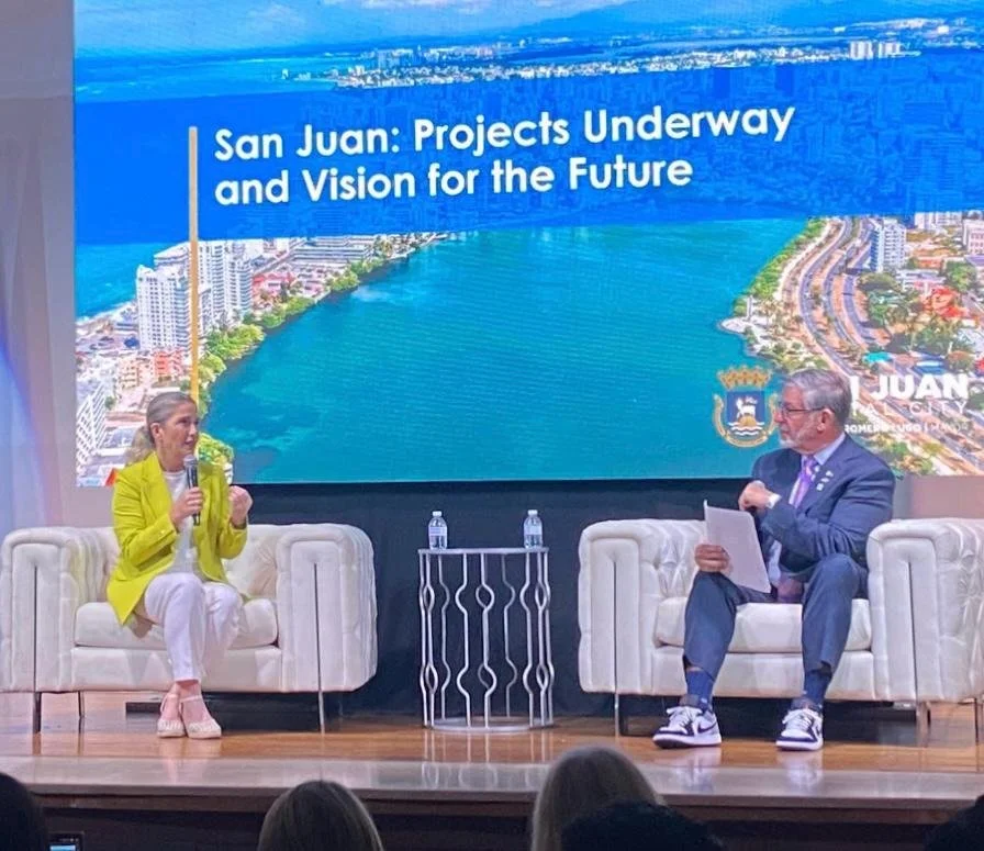 A woman and a man sit on stage in chairs, engaged in a discussion. The woman wears a yellow blazer, white pants, and pink shoes. The man wears a suit, sneakers, and glasses. Behind them, a large screen displays a photo of San Juan with the title 'San Juan: Projects Underway and Vision for the Future'.