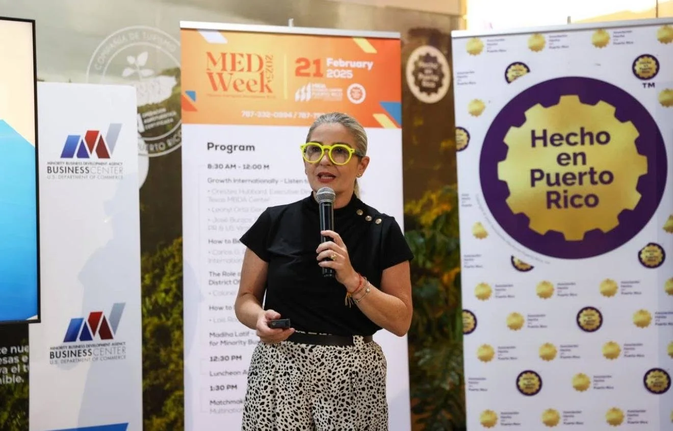 A woman with gray hair, yellow glasses, and a black shirt speaking into a microphone at a conference. Behind her are banners, including one that says "Hecho en Puerto Rico" and another for MED Week 2025.