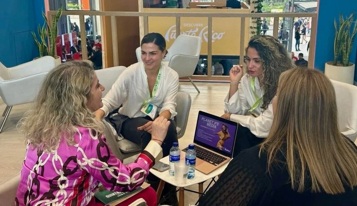 Four women sitting in a circle at a table engaged in conversation at a conference or meeting. There are water bottles and a laptop on the table, with an indoor setting and people visible outside through large windows.