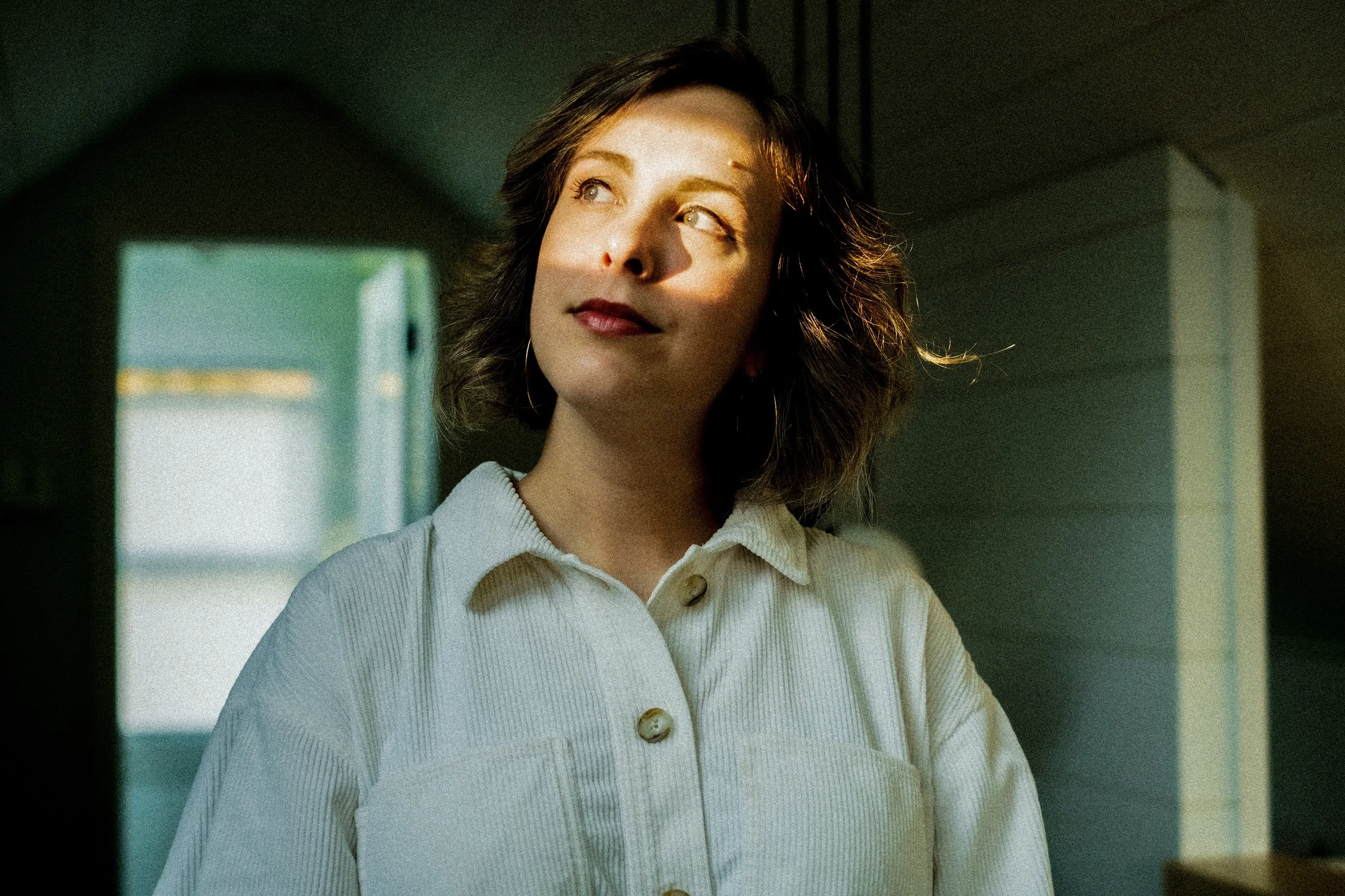 A woman with short wavy brown hair and light makeup looking away, with sunlight on her face, wearing a light-colored button-up shirt in a dimly lit room.
