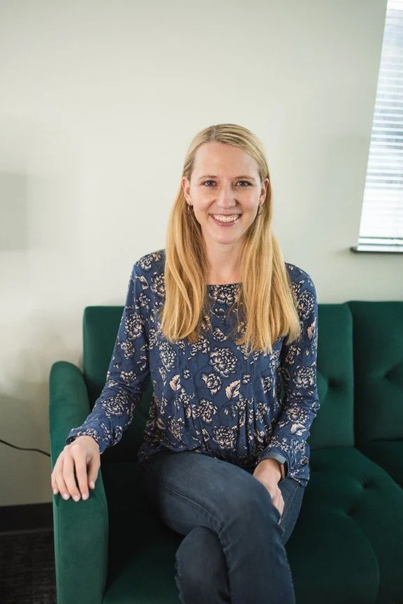A woman with long blonde hair, wearing a blue floral blouse, sitting on a green couch in a room with light-colored walls and window blinds.