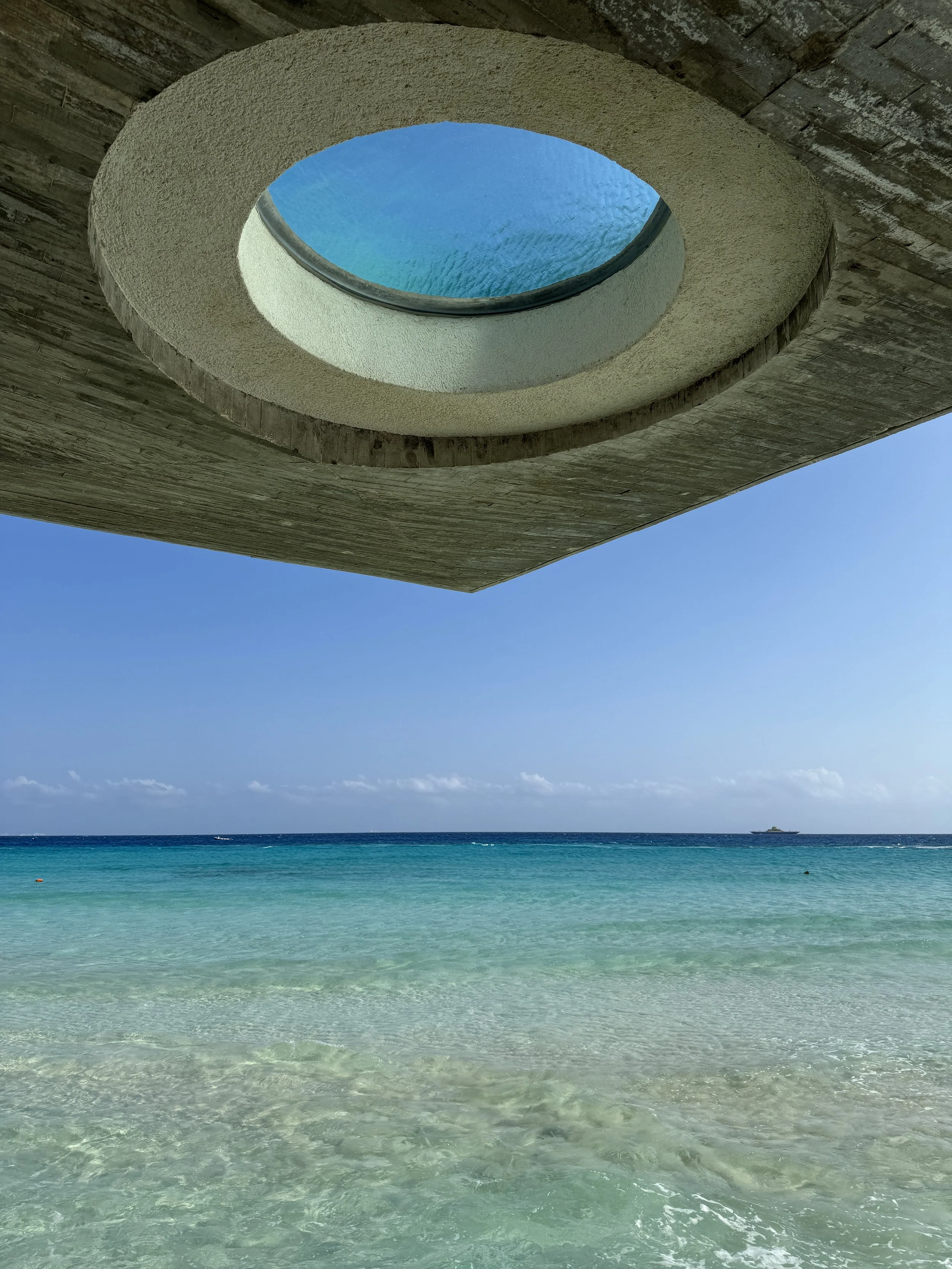 Looking up through a circular opening in a concrete structure to view the blue sky and ocean. The ocean water is clear and turquoise near the shore, with a boat in the distance.