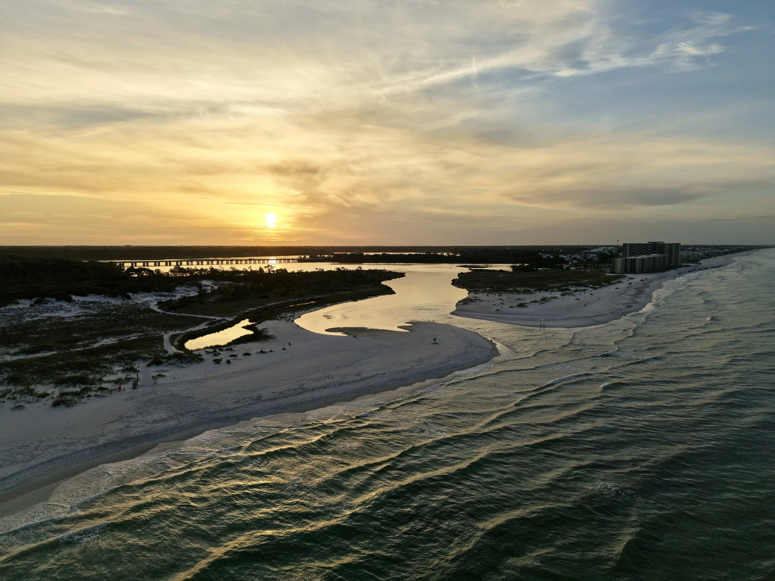 Sunset over a river with a bridge, sandy beach, water, and a city skyline in the distance.
