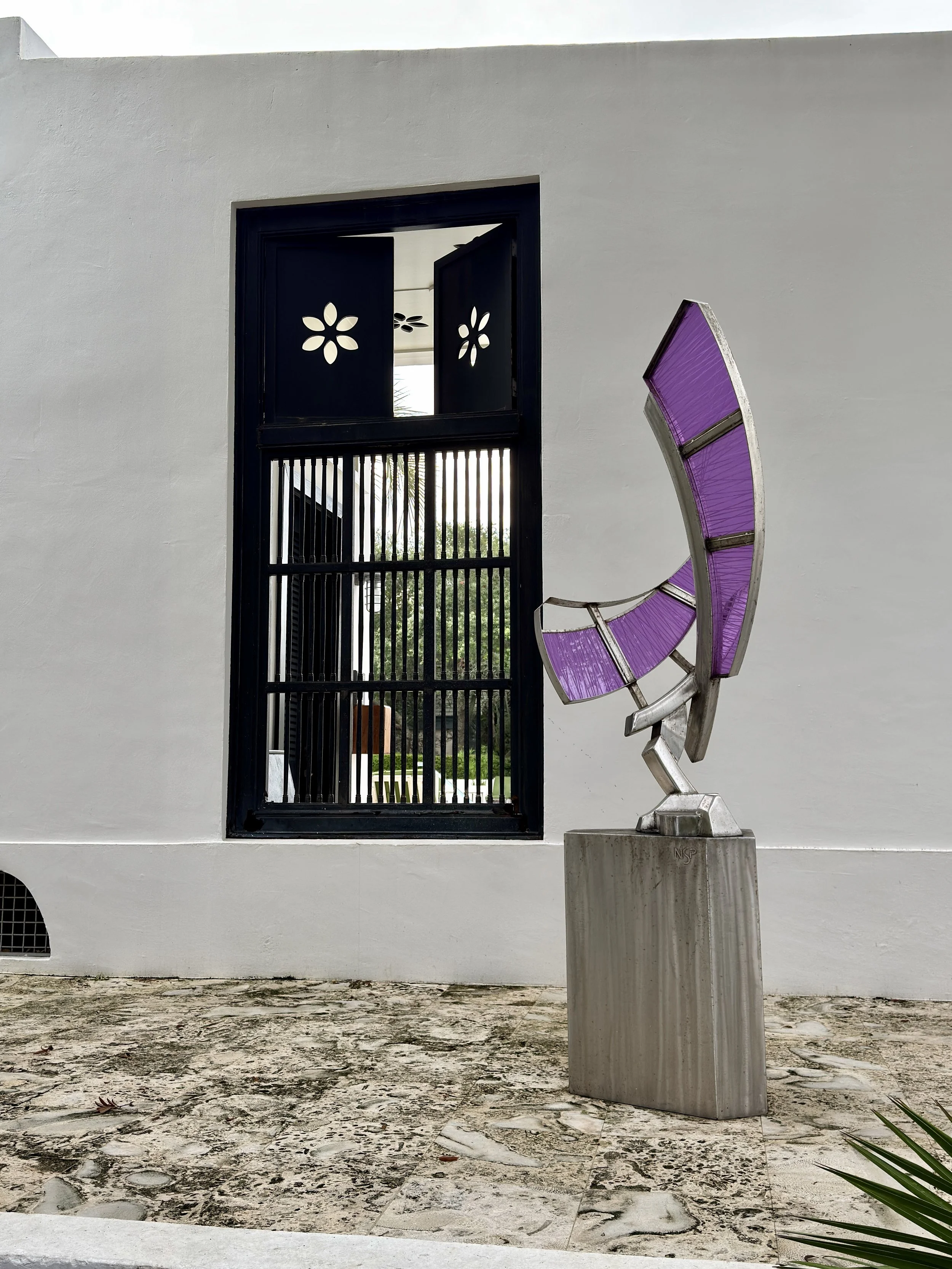 Modern sculpture with purple geometric panels mounted on a silver metallic structure, situated on a concrete base in front of a white building with a black metal window with decorative cutouts.