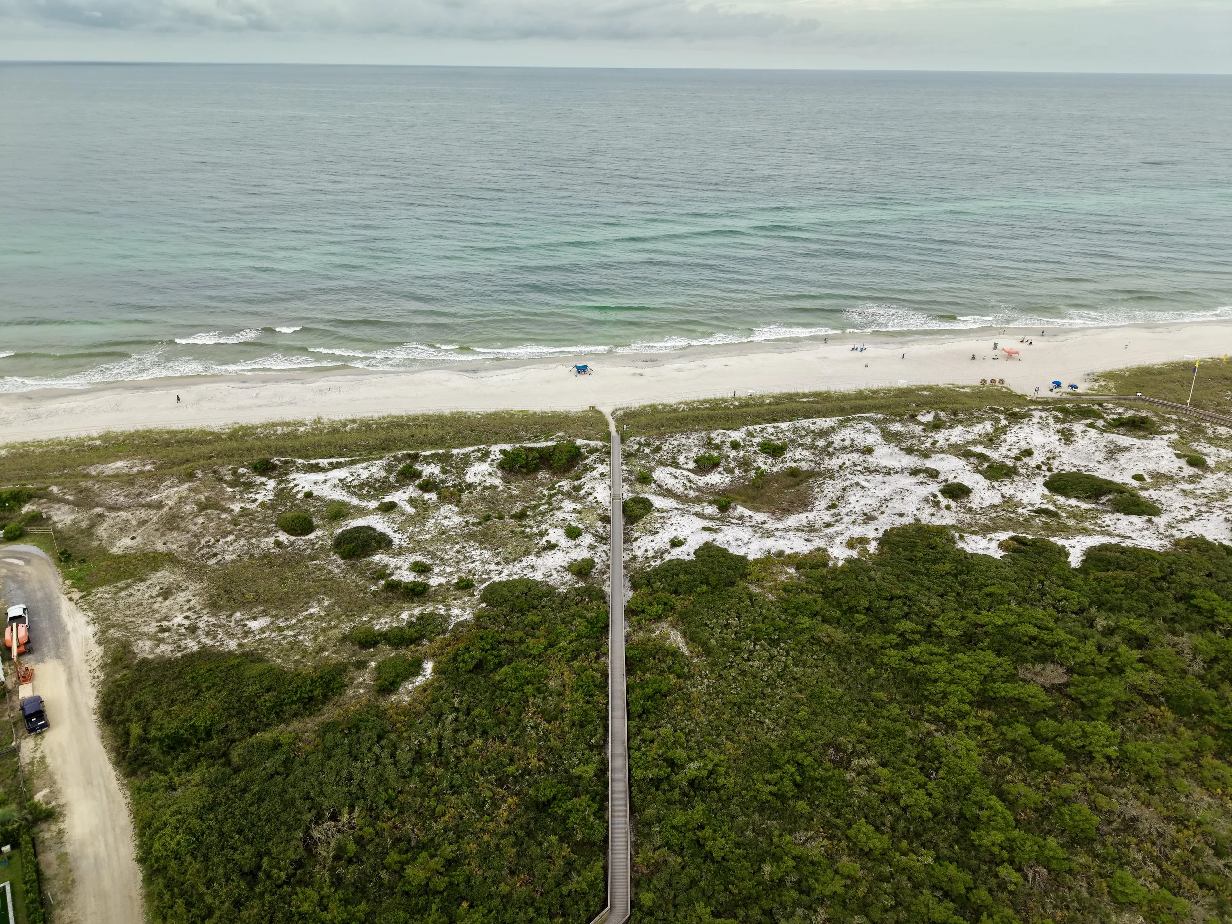 Aerial view of a beach with white sand and greenish-blue water, lined with green bushes and a wooden walkway.