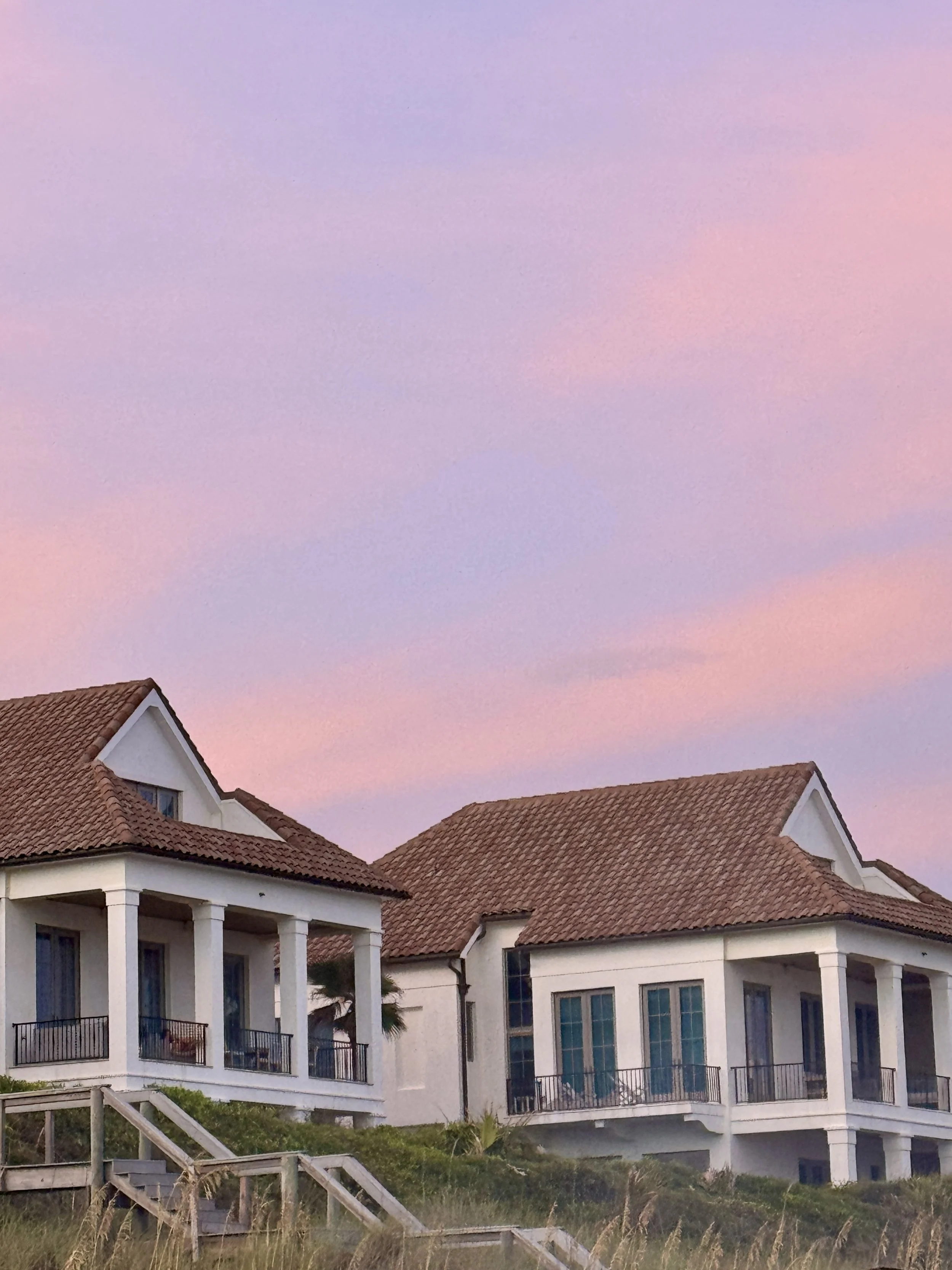 A multi-story white house with red-tiled roofs, large windows, and balconies, situated on a grassy hill with a wooden staircase leading up to it, under a pastel-colored sky at dusk.