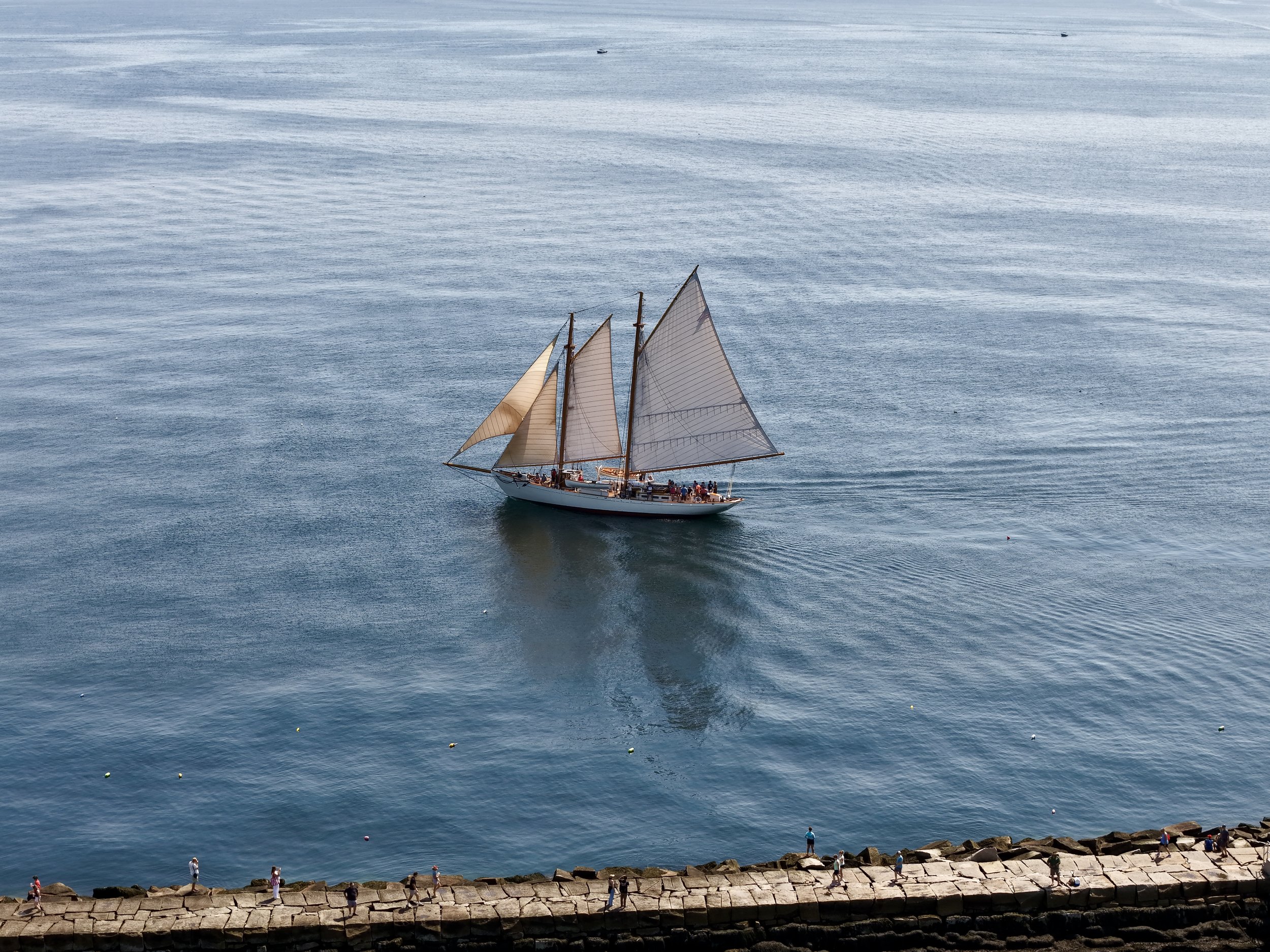 A sailboat with multiple sails on a calm ocean, viewed from the shoreline with people walking nearby.
