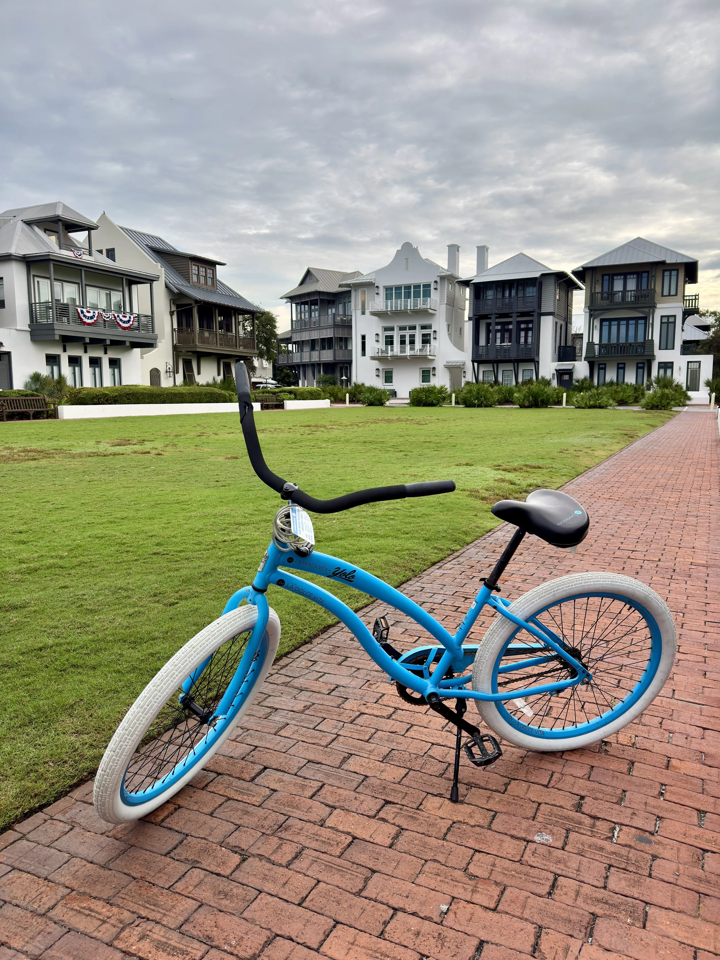 A blue bicycle with white tires on a brick pathway in a park area, with modern residential buildings in the background under a cloudy sky.