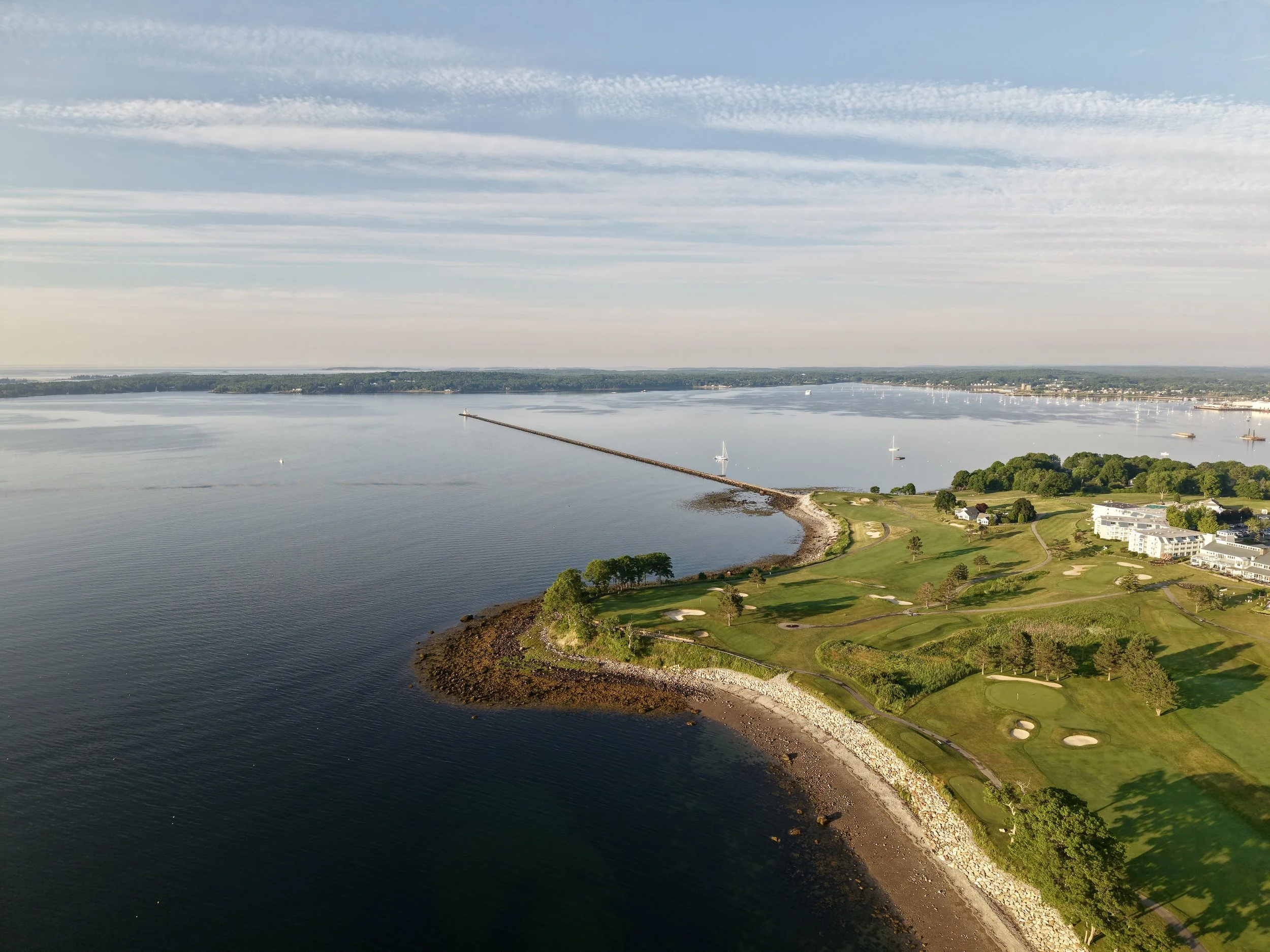 Aerial view of a large body of water with sailboats, a shoreline with a golf course and residential buildings, and a breakwater extending into the water.