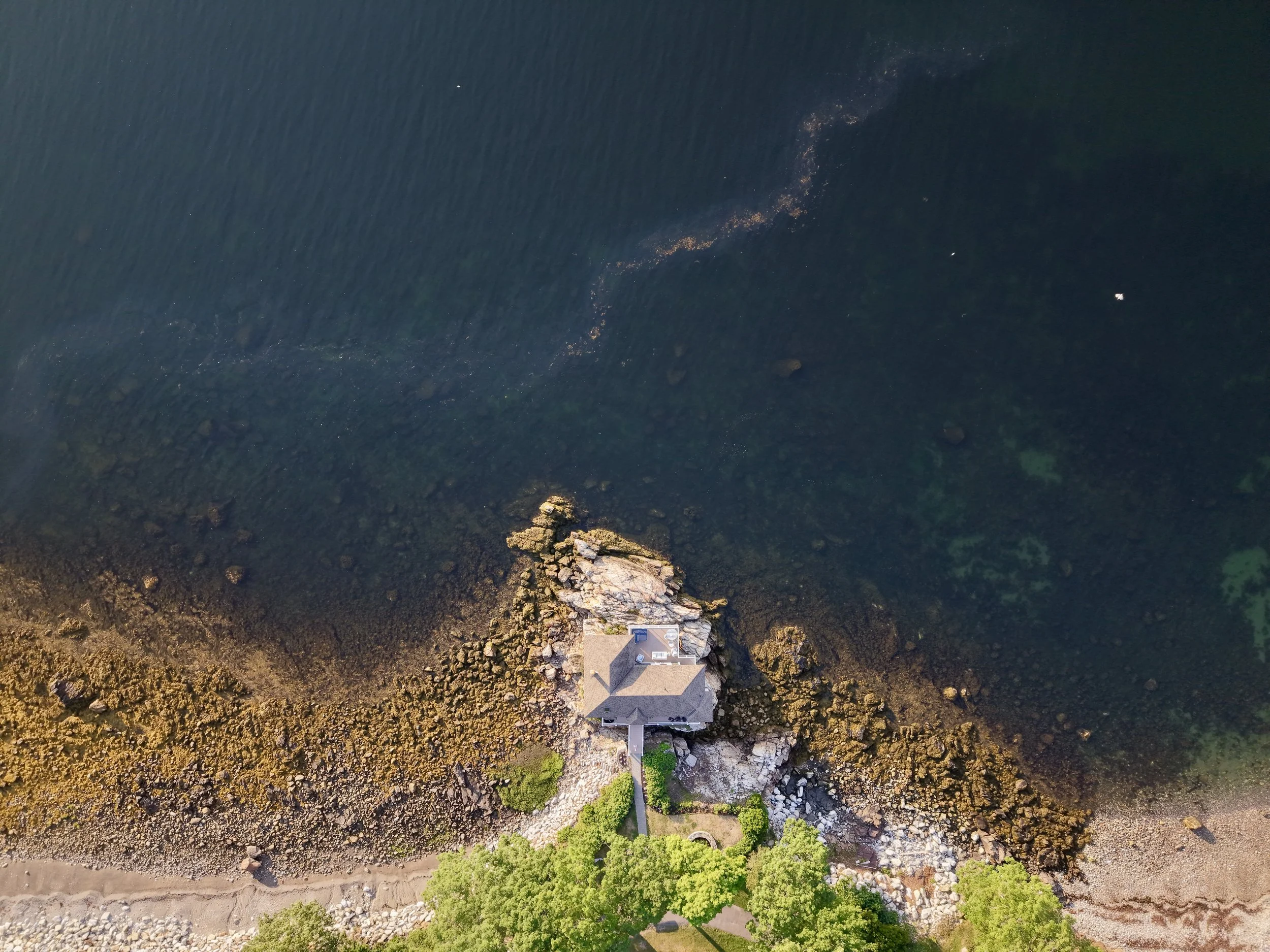 Aerial view of a house on rocky terrain with a patch of green grass, near a body of water, with rocks and trees visible around.