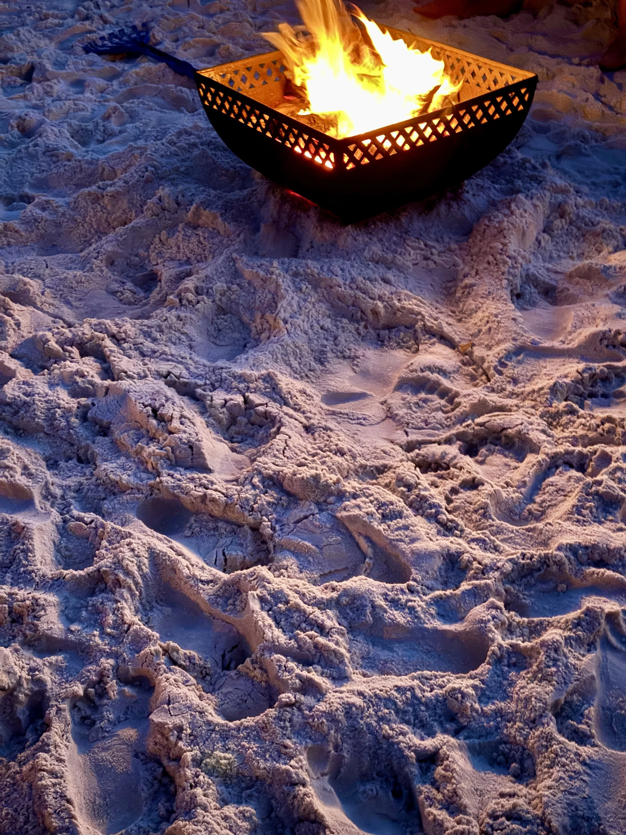 A fire burning in a black metal fire pit on snow, with footprints and churned snow around it, during twilight or early evening.