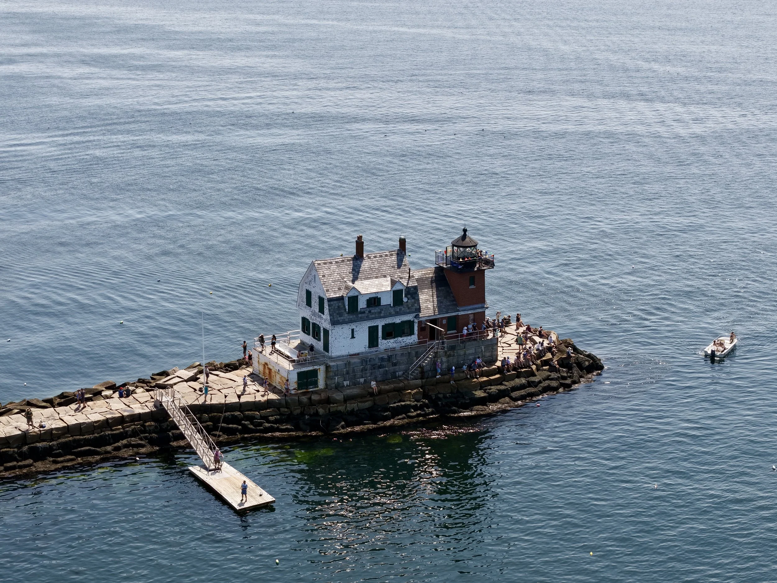 A lighthouse building with a red roof and a rooftop observation deck, located on a rocky shore, with many people gathering around it and walking along the pier, overlooking a large body of water with gentle waves.