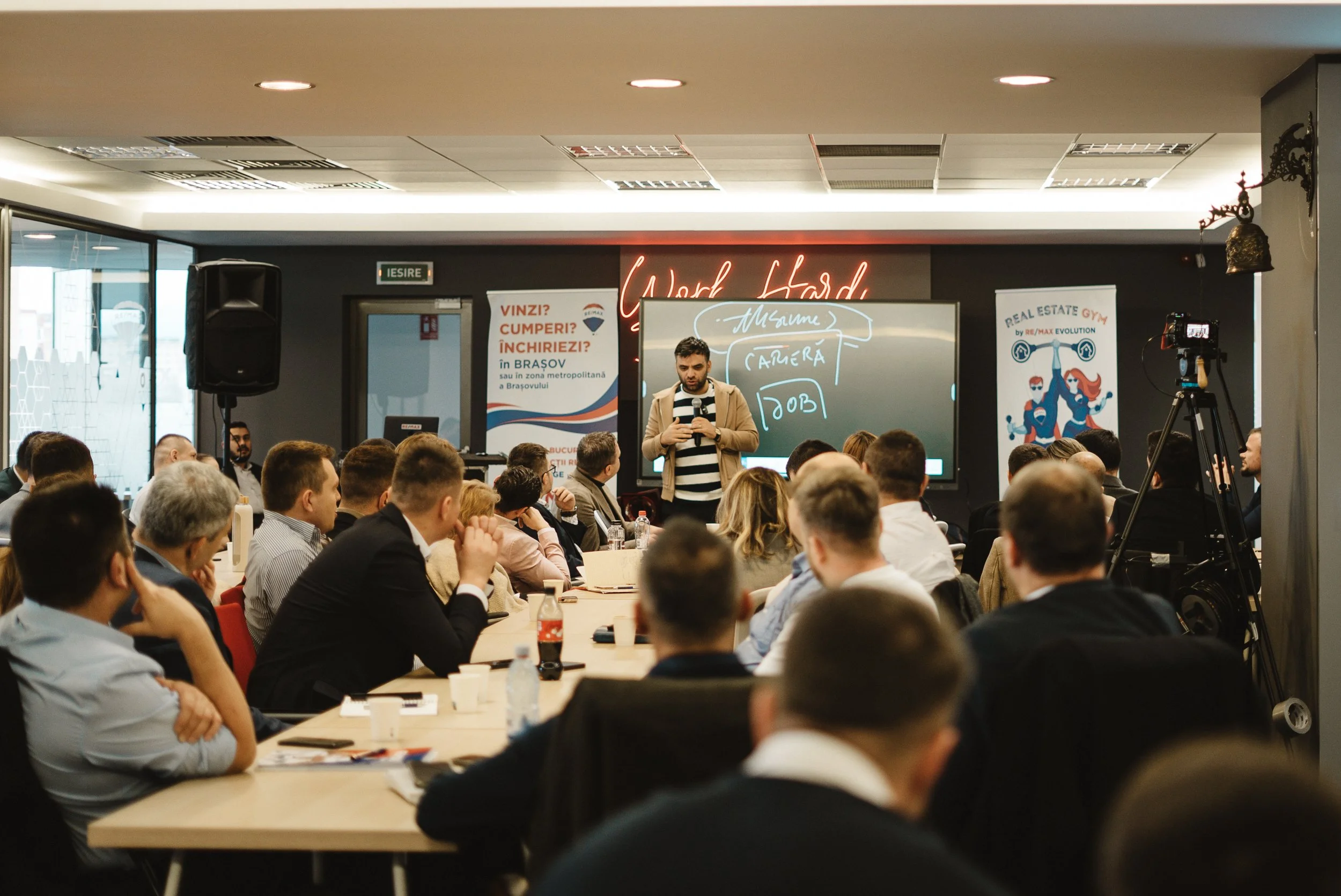 A man giving a presentation to a group of people in a conference room with a blackboard and cameras.