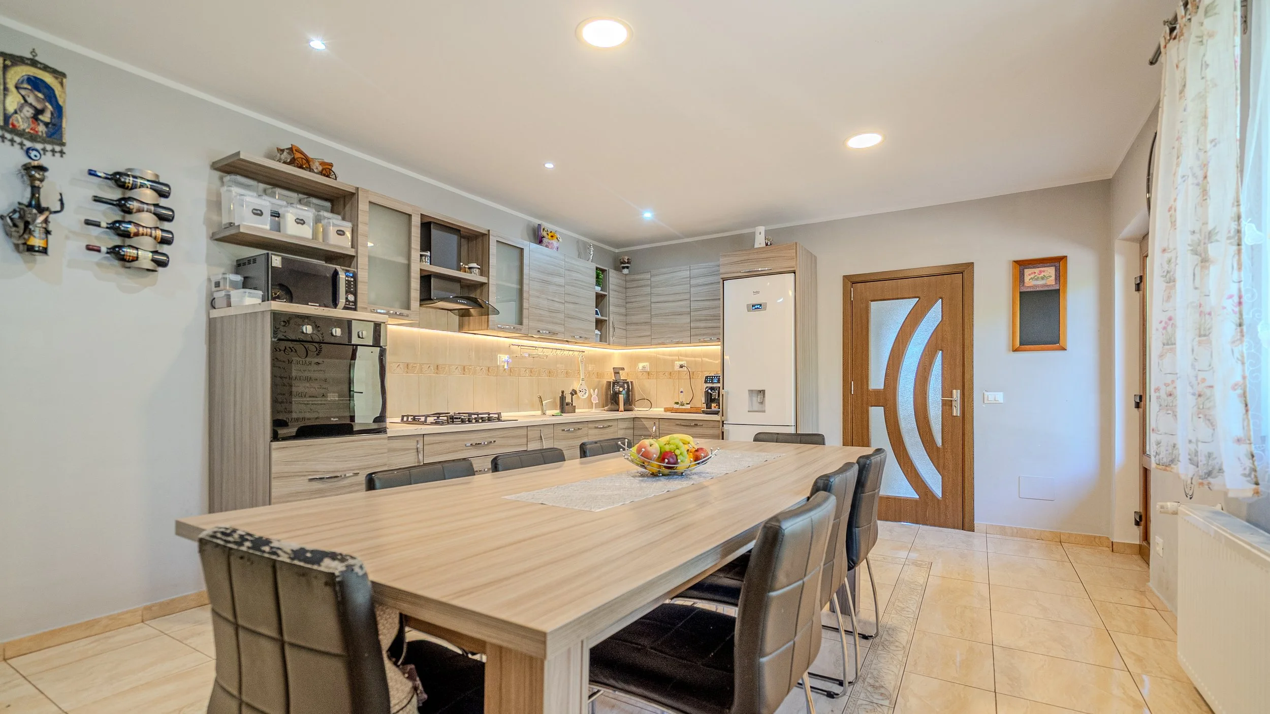 Kitchen with a wooden dining table and black chairs, light wood cabinets, a door with a circular design, and a window with floral curtains.