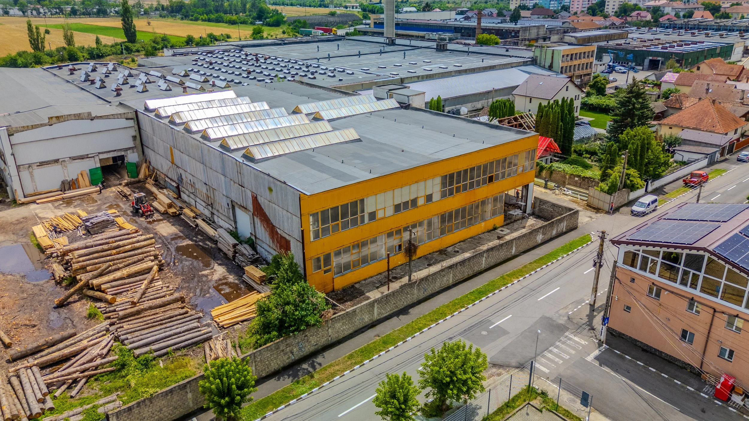 Aerial view of a construction site next to a yellow building with large windows, piles of logs, a forklift, and neighboring houses with gardens and solar panels.