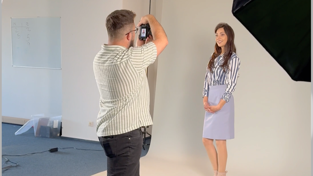 A woman posing for a photo in front of a white backdrop while a man takes her picture with a camera, in a photo studio setting.