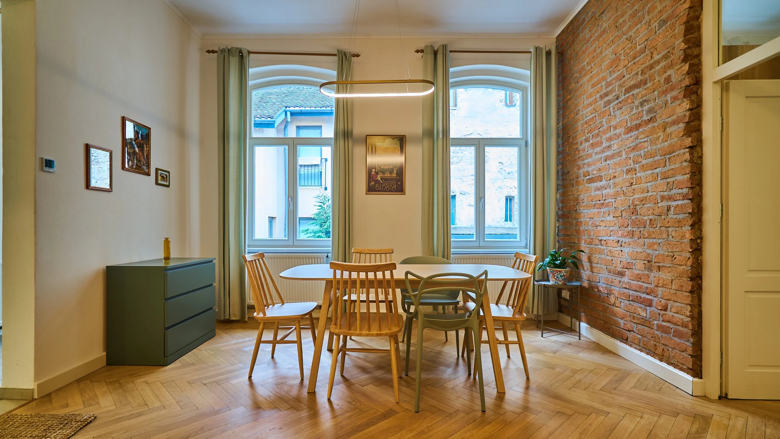 Dining room with wooden floor, round dining table surrounded by mixed wooden and plastic chairs, brick accent wall, two large windows with green curtains, and a small side table with a potted plant.
