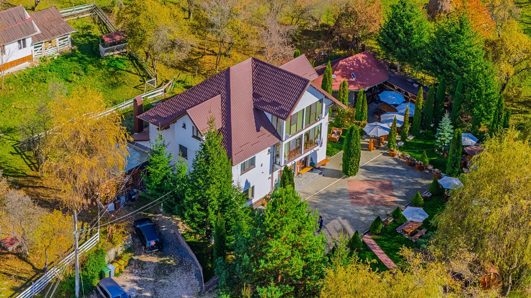 Aerial view of a large house with a red roof surrounded by trees with autumn foliage, a paved courtyard, and outdoor seating under umbrellas.