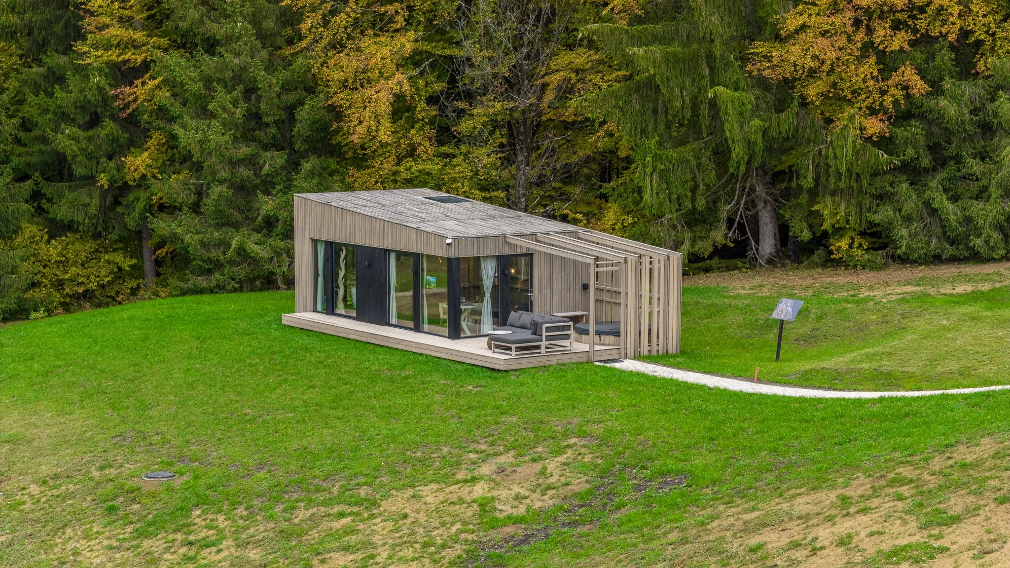 A modern small house with large glass doors is situated on a grassy area, with a dense forest in the background. The house has a wooden exterior and an outdoor deck with outdoor furniture. There is a signboard on a pole near the house.