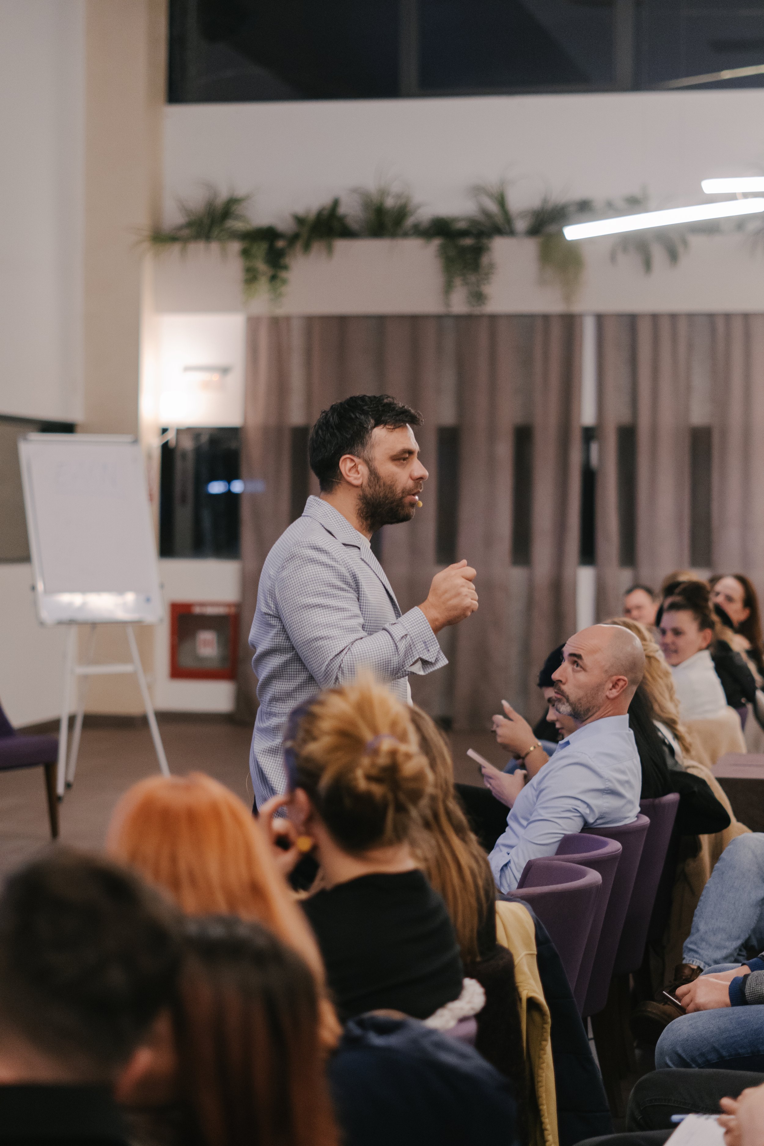A man in a blazer giving a presentation at a conference or seminar, with an audience seated and listening attentively.