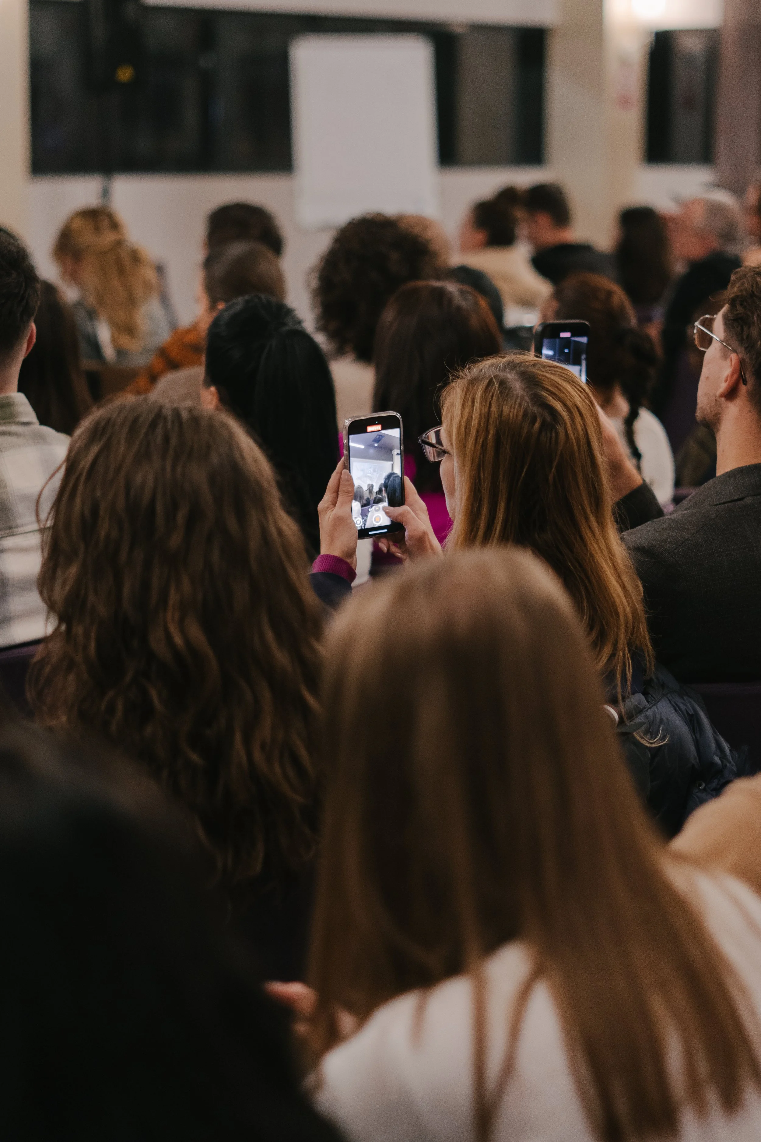 A group of people attending a presentation or conference, with some taking pictures using their smartphones.