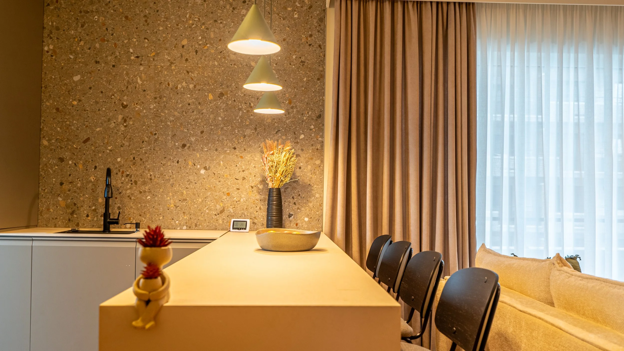 Modern kitchen and dining area with beige walls, a countertop with a black faucet, a vase with dried flowers, a bowl, and a small clock, next to a set of black chairs, beige curtains, and a beige sofa.