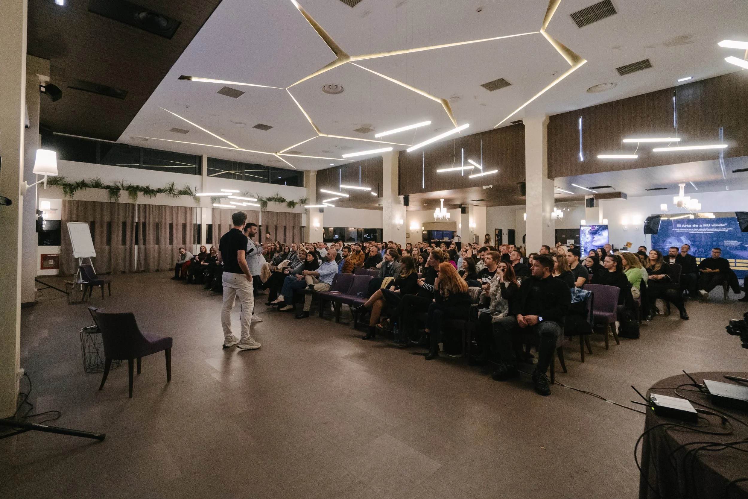 A large group of people seated in an auditorium listening to two presenters standing in front of the audience, with a large screen displaying a presentation in the background.