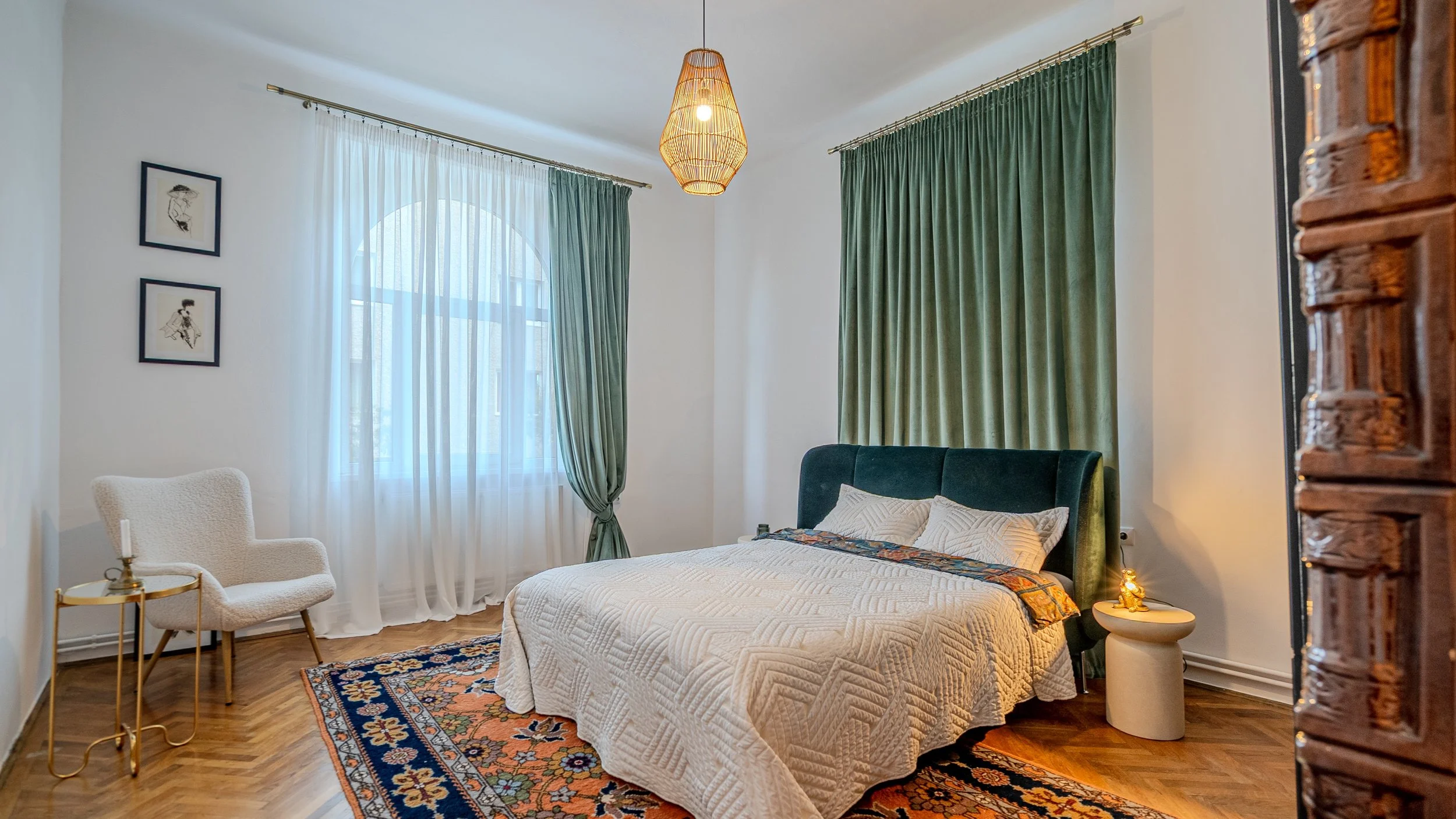 A bedroom with a white quilted bedspread, two pillows, and a dark green velvet headboard. There are two windows with sheer white and dark green curtains, a white armchair with wooden legs and a gold side table with a candle, and a colorful patterned 