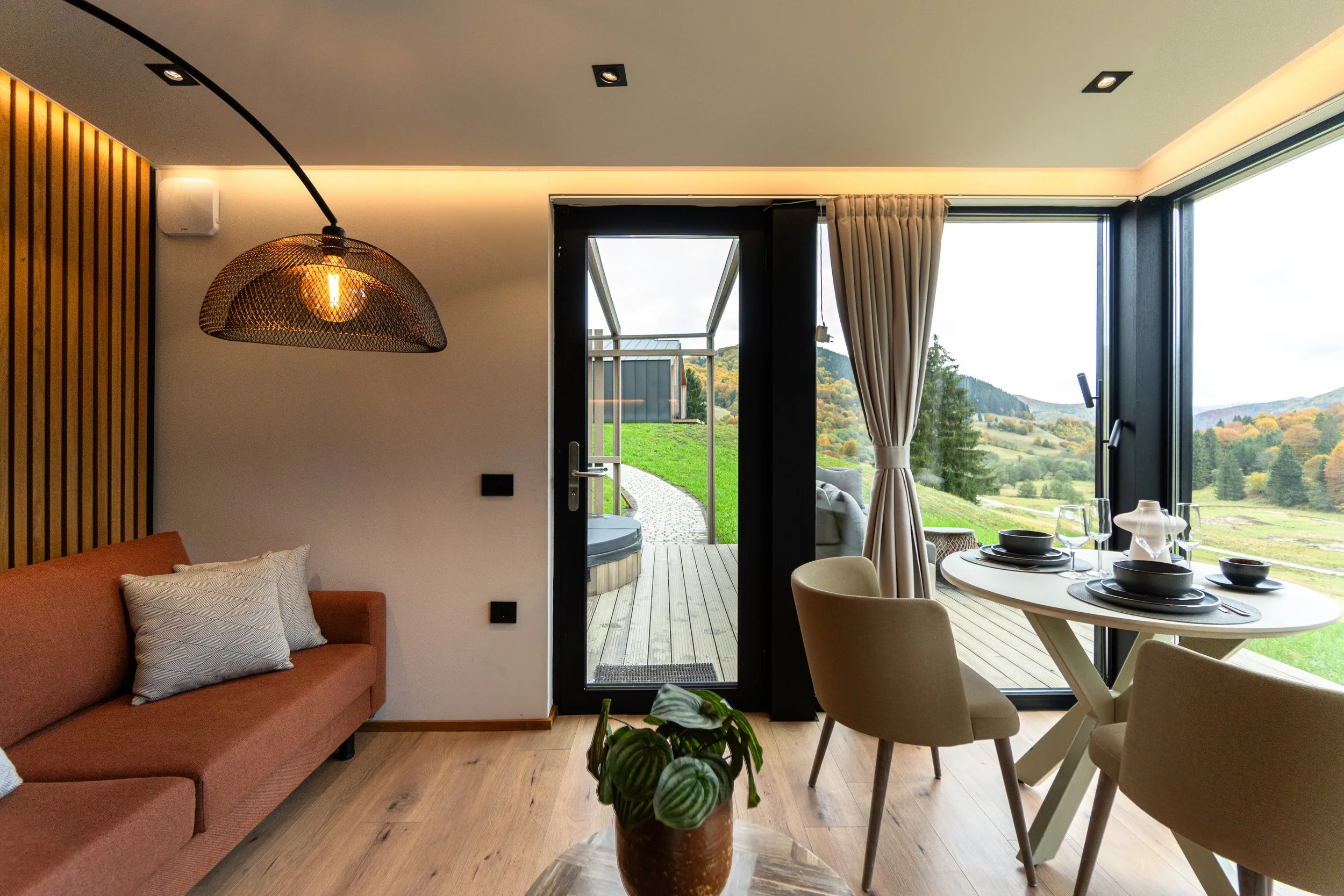 Living room with a brown couch, a hanging lamp, a round dining table set with plates, glasses, and a napkin, overlooking a scenic outdoor landscape with green hills and trees through large windows and a door leading to a deck.
