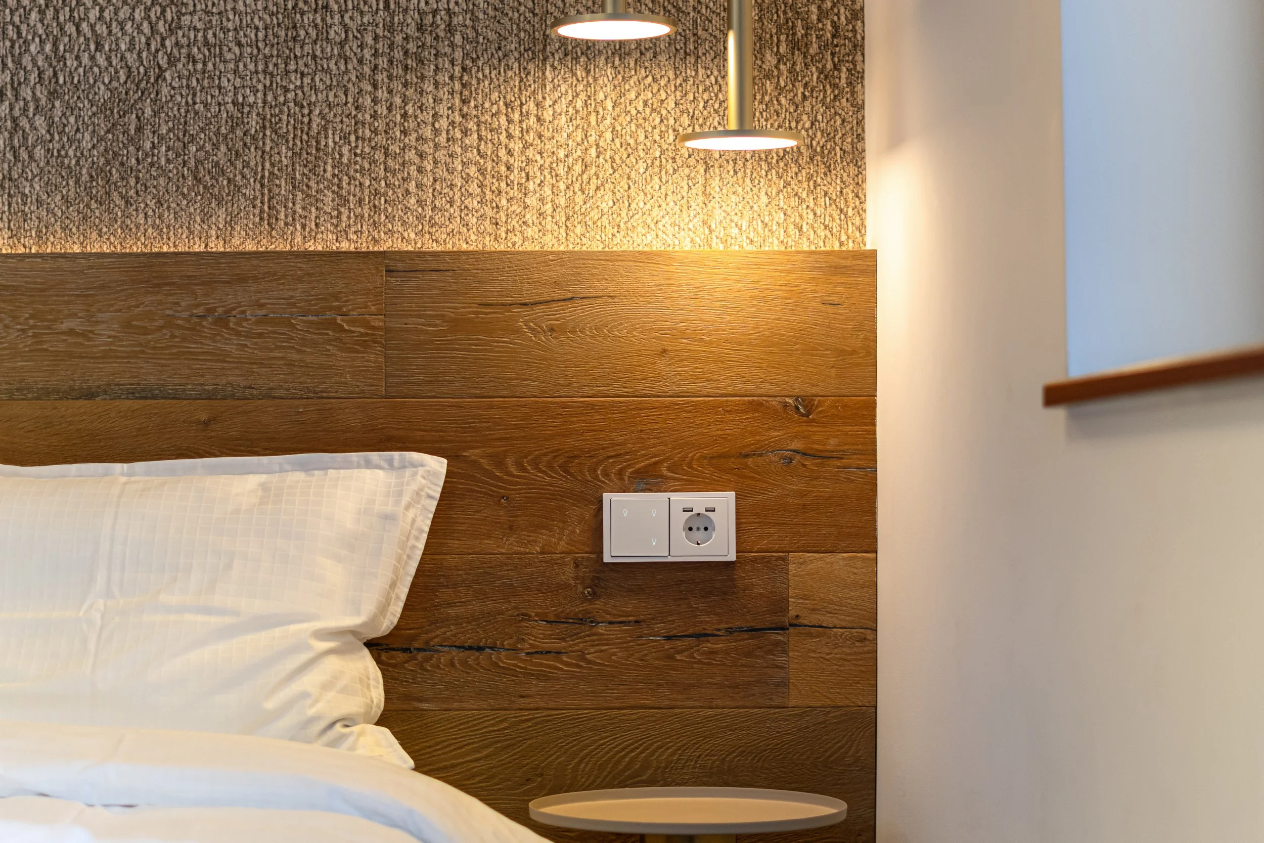 Close-up of a bedroom headboard with a bedside table, electrical outlets, and a wall-mounted lamp, with textured wall behind.