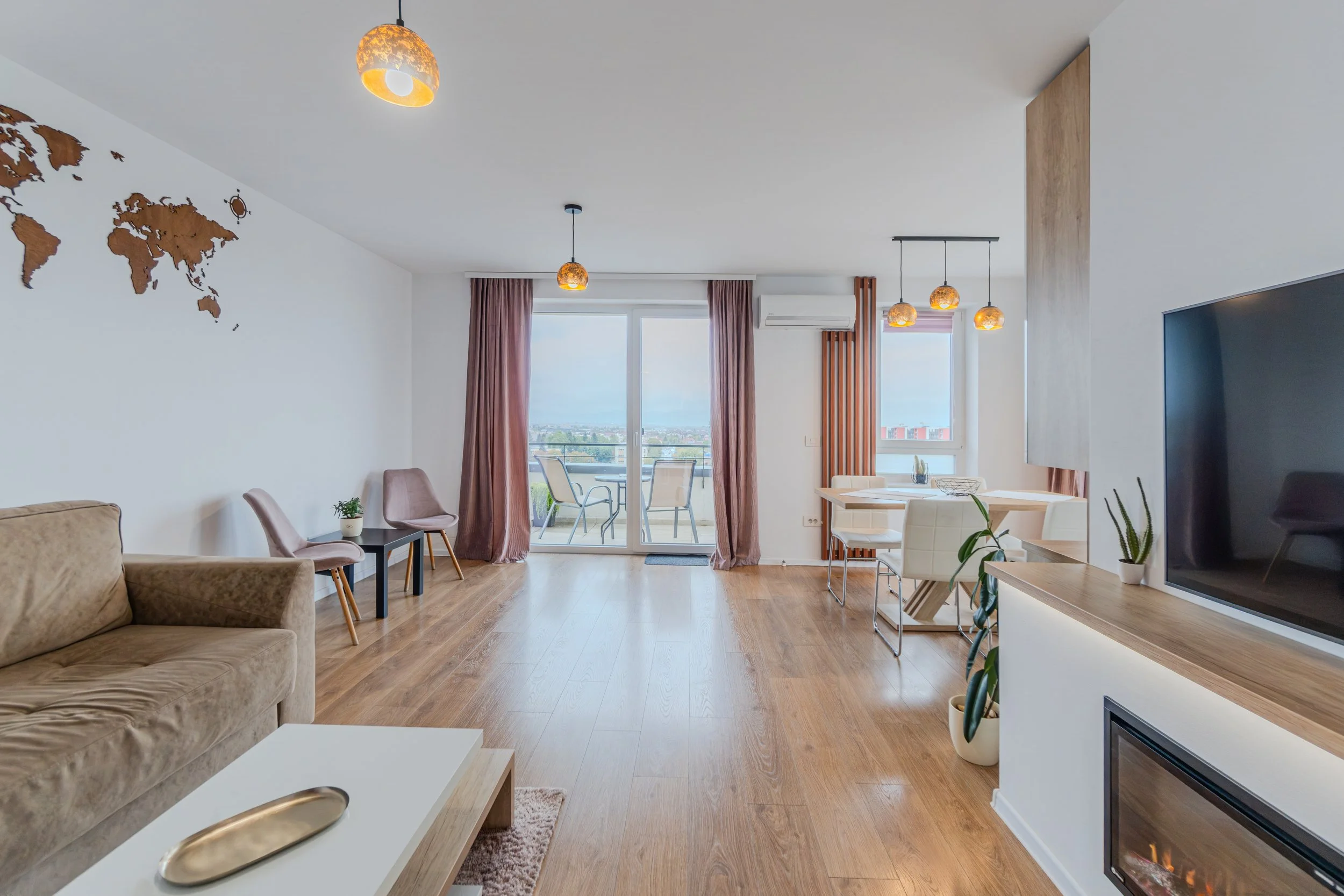 Living room with wooden flooring, beige sofa, pink chairs, a white coffee table with a golden tray, a flat-screen TV and a fireplace, a dining area with white chairs and a wooden table, sliding glass doors with pink curtains leading to a balcony, and