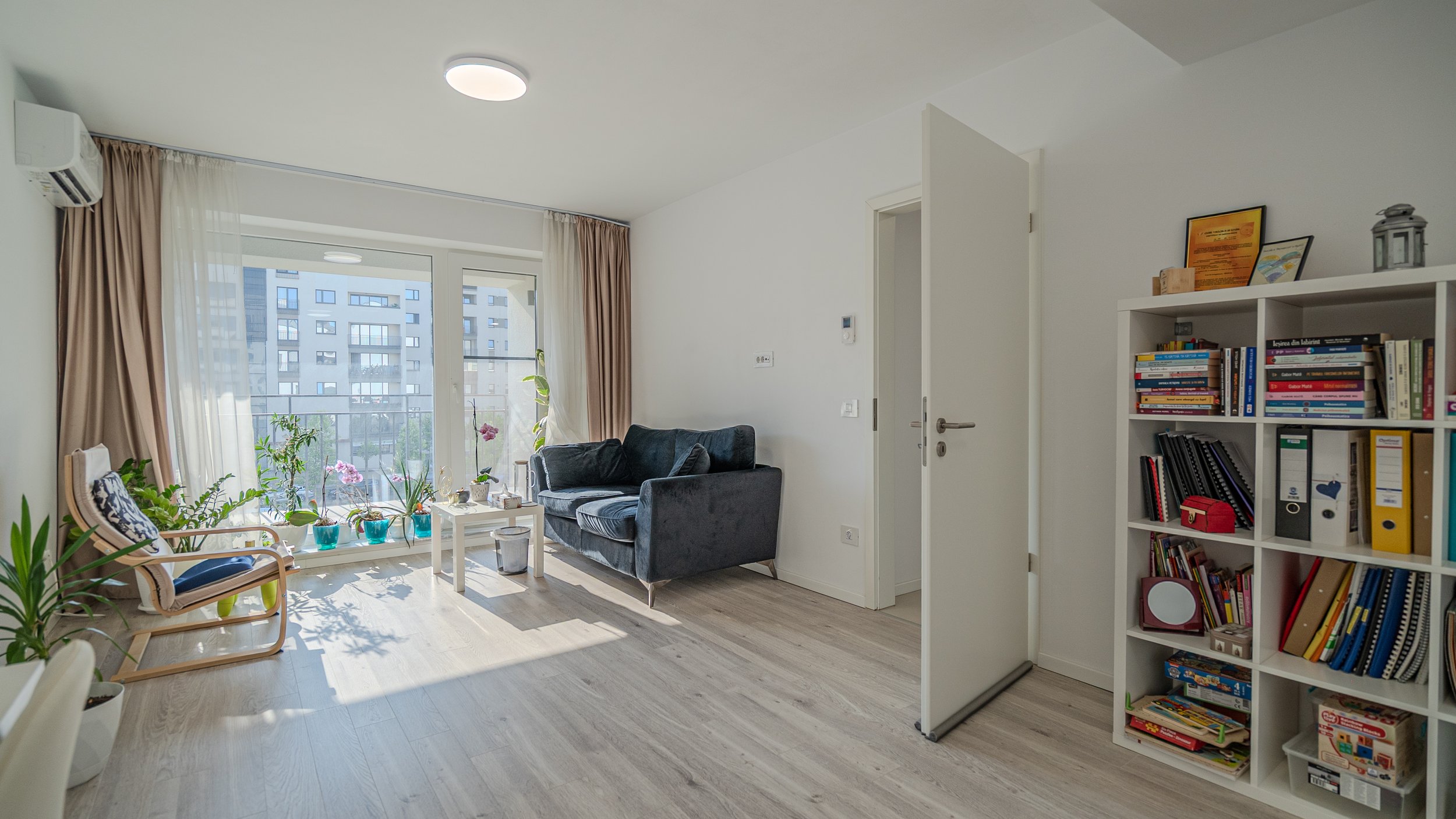 Bright living room with large windows, beige curtains, a black sofa, wooden armchair, white coffee table, bookshelf with books and decorative items, and potted plants on the floor and windowsill.