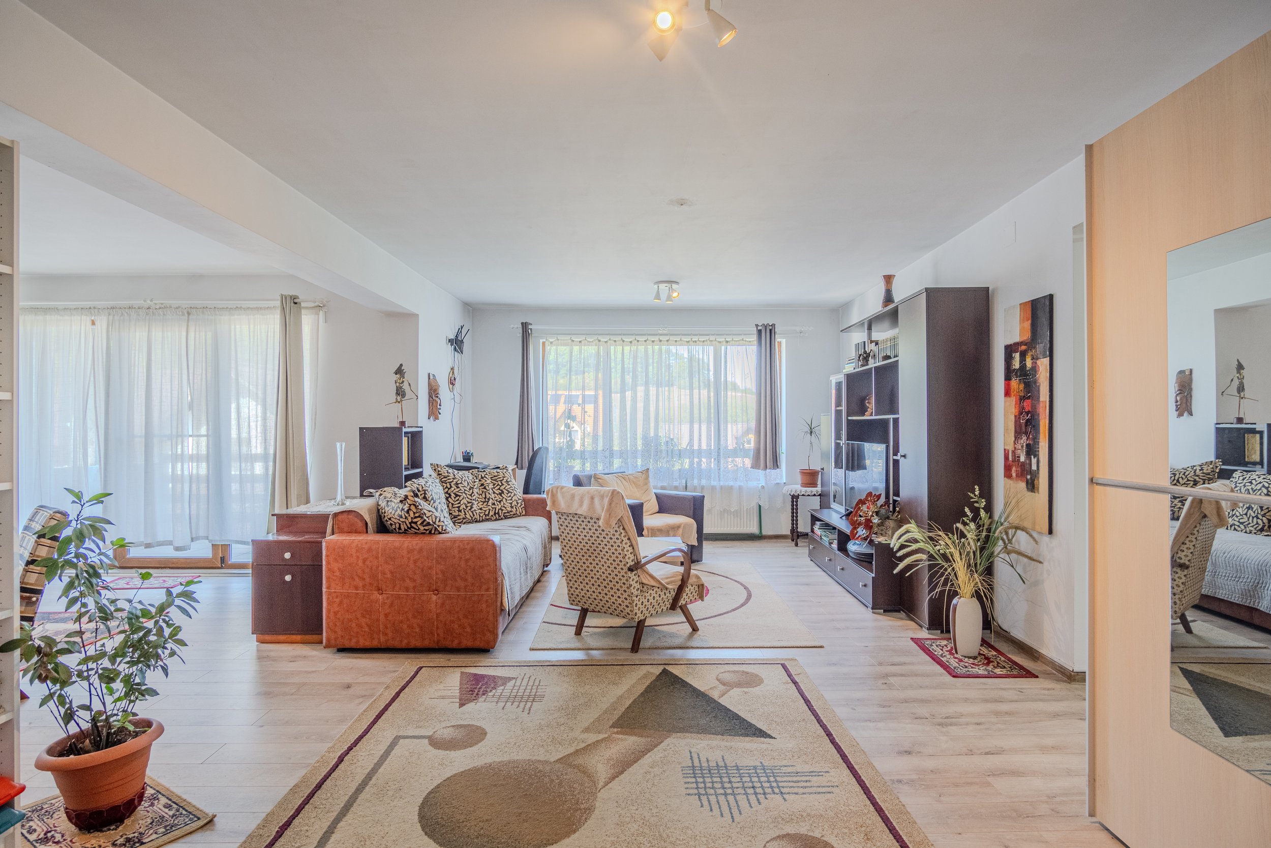 Bright living room with large windows, orange sofa, patterned chairs, TV, bookshelf, indoor plants, and decorative wall art.