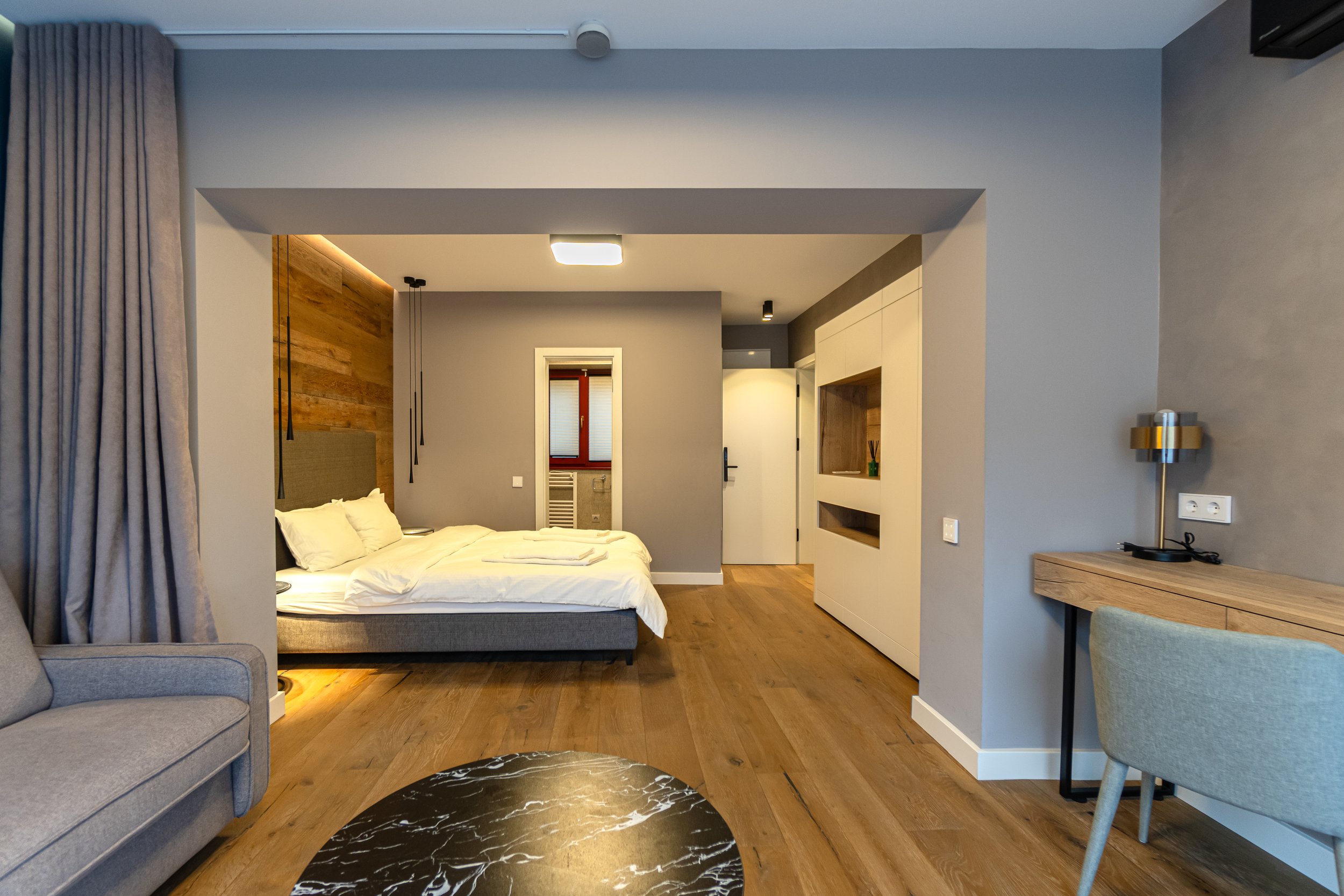 Modern hotel room with a bed, wooden accent wall, and a sitting area with a gray sofa and a round black marble table.
