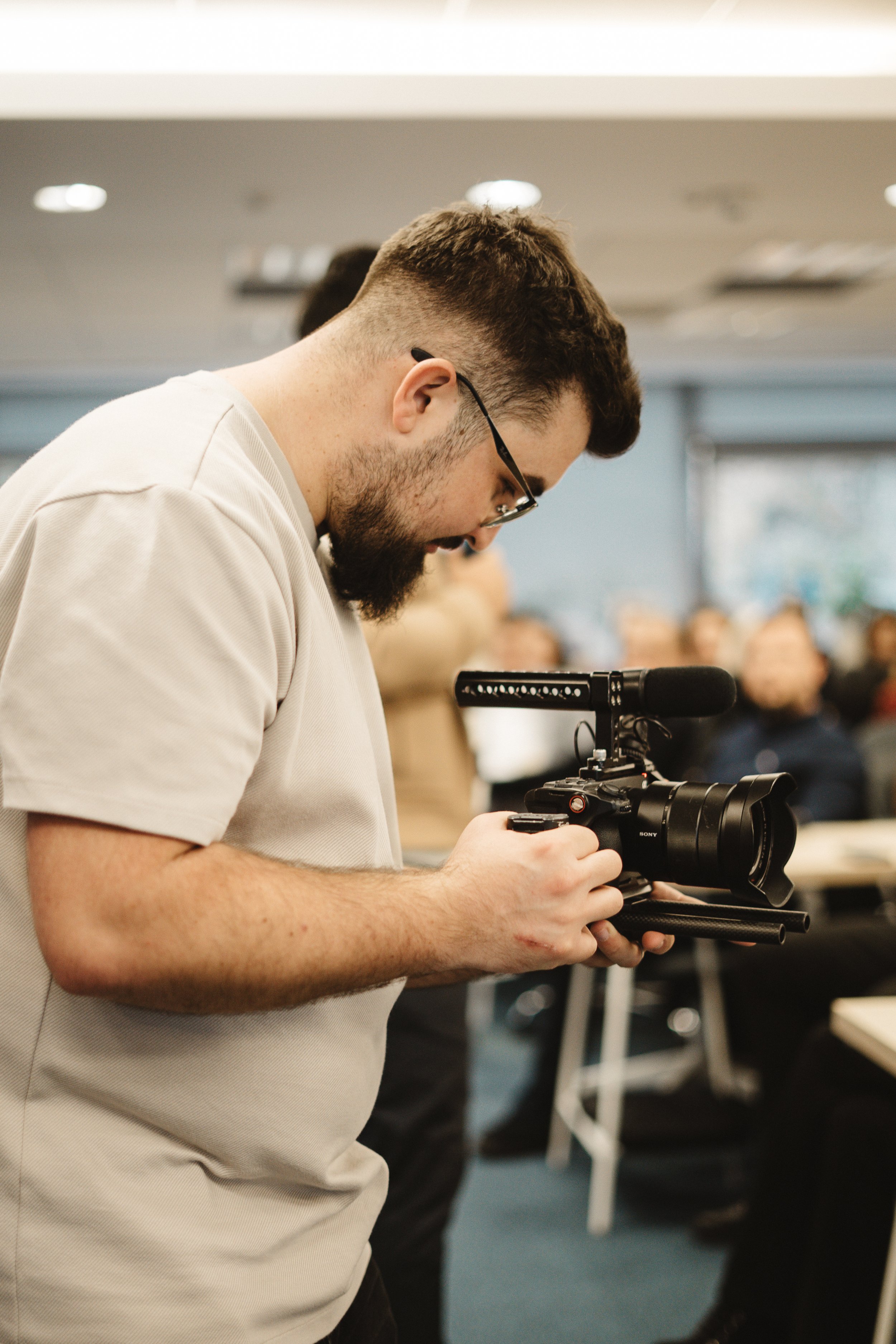 A man with glasses and a beard is operating a professional video camera in a room filled with people.