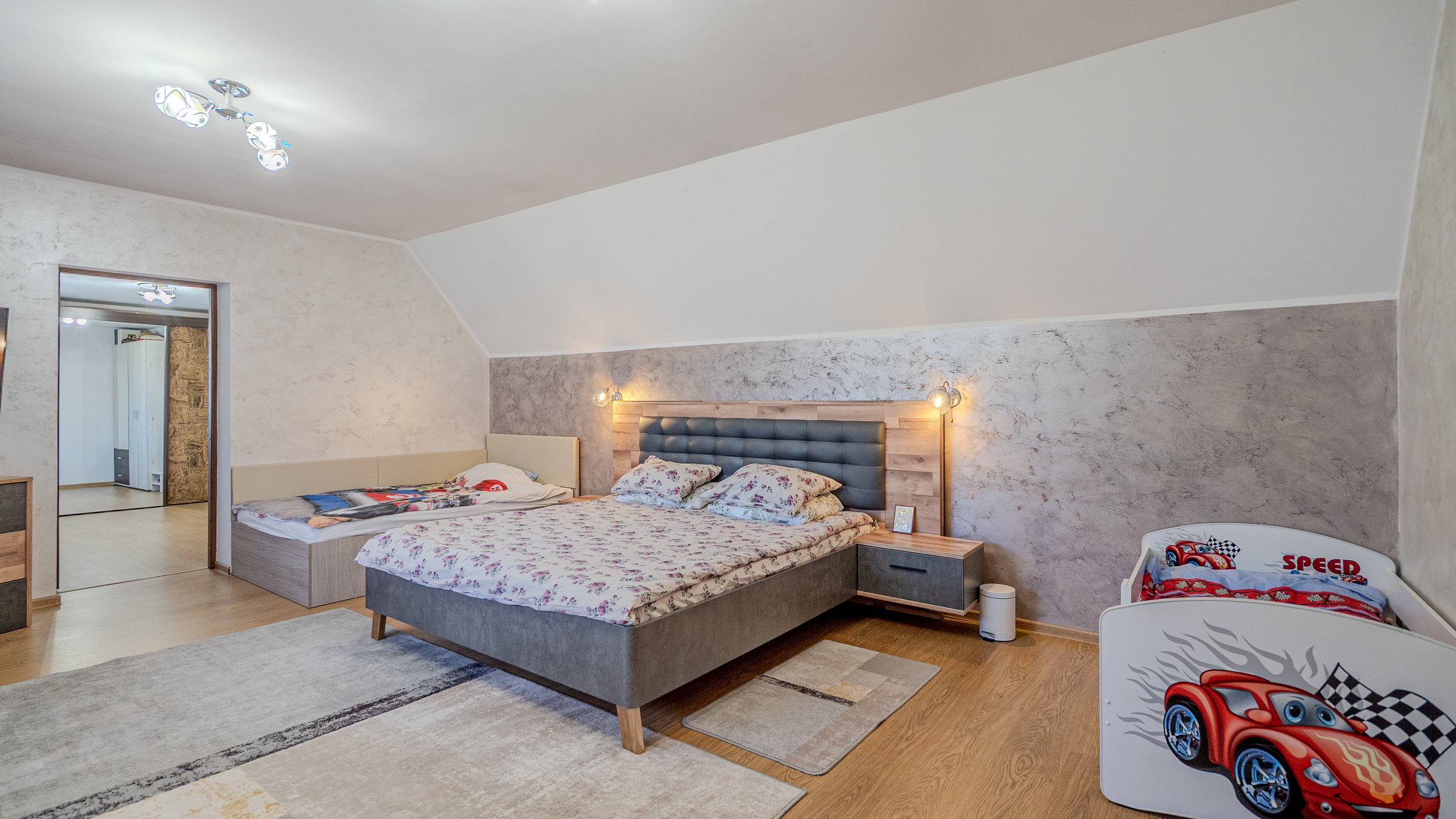 A bedroom with a large bed, a nightstand, and a small children's bed with a race car motif. The room has light-colored walls, a patterned wall behind the large bed, and wooden flooring.
