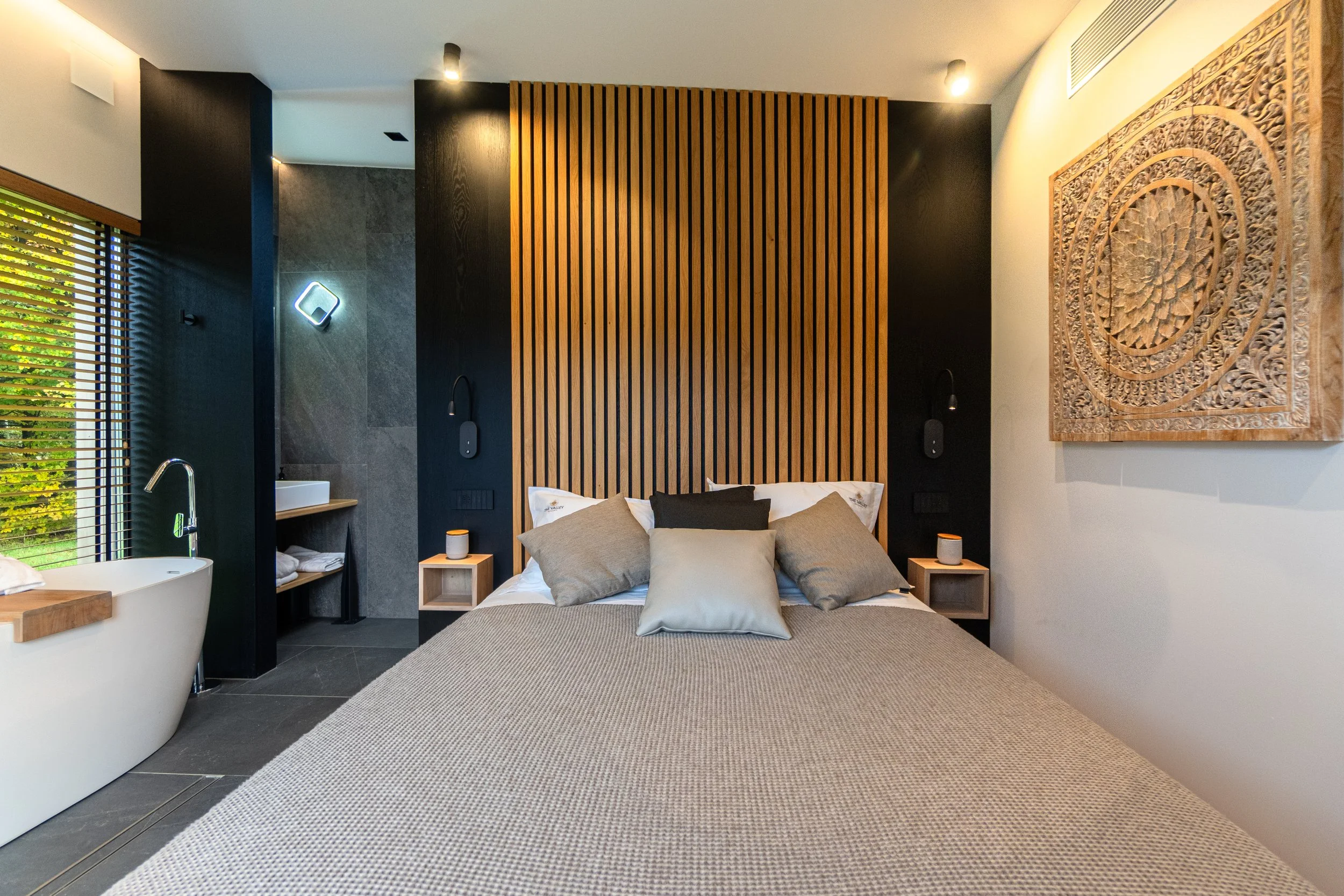 Modern bedroom with large bed, decorative wooden headboard, and neutral-colored bedding. To the left, a bathtub near a window with blinds. The wall behind the bed features dark and wood-paneled sections with wall-mounted reading lights. There's a sma