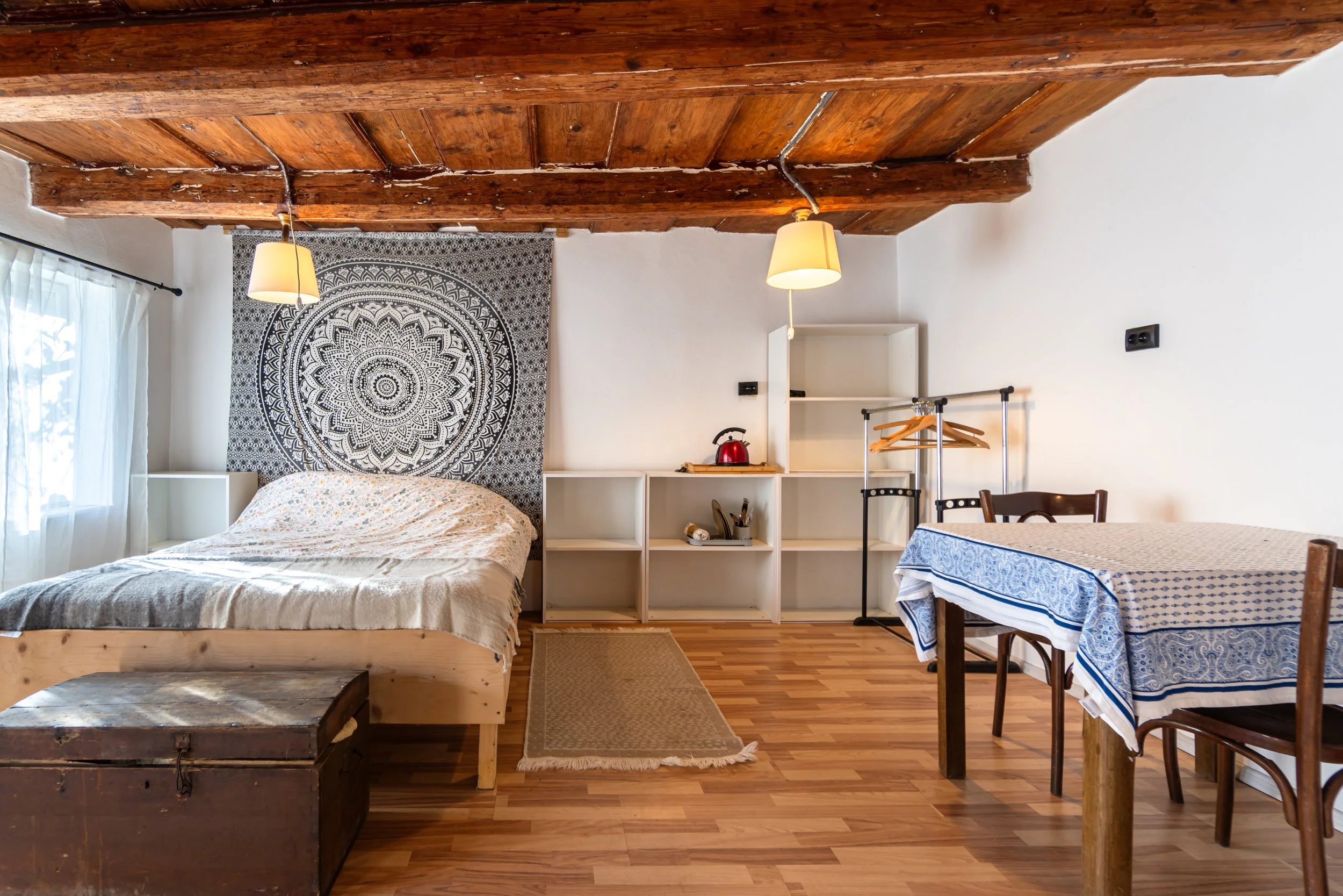 Cozy bedroom with wooden ceiling, tapestry behind bed, white shelving, table with blue patterned tablecloth, chairs, and a storage chest at the foot of the bed.