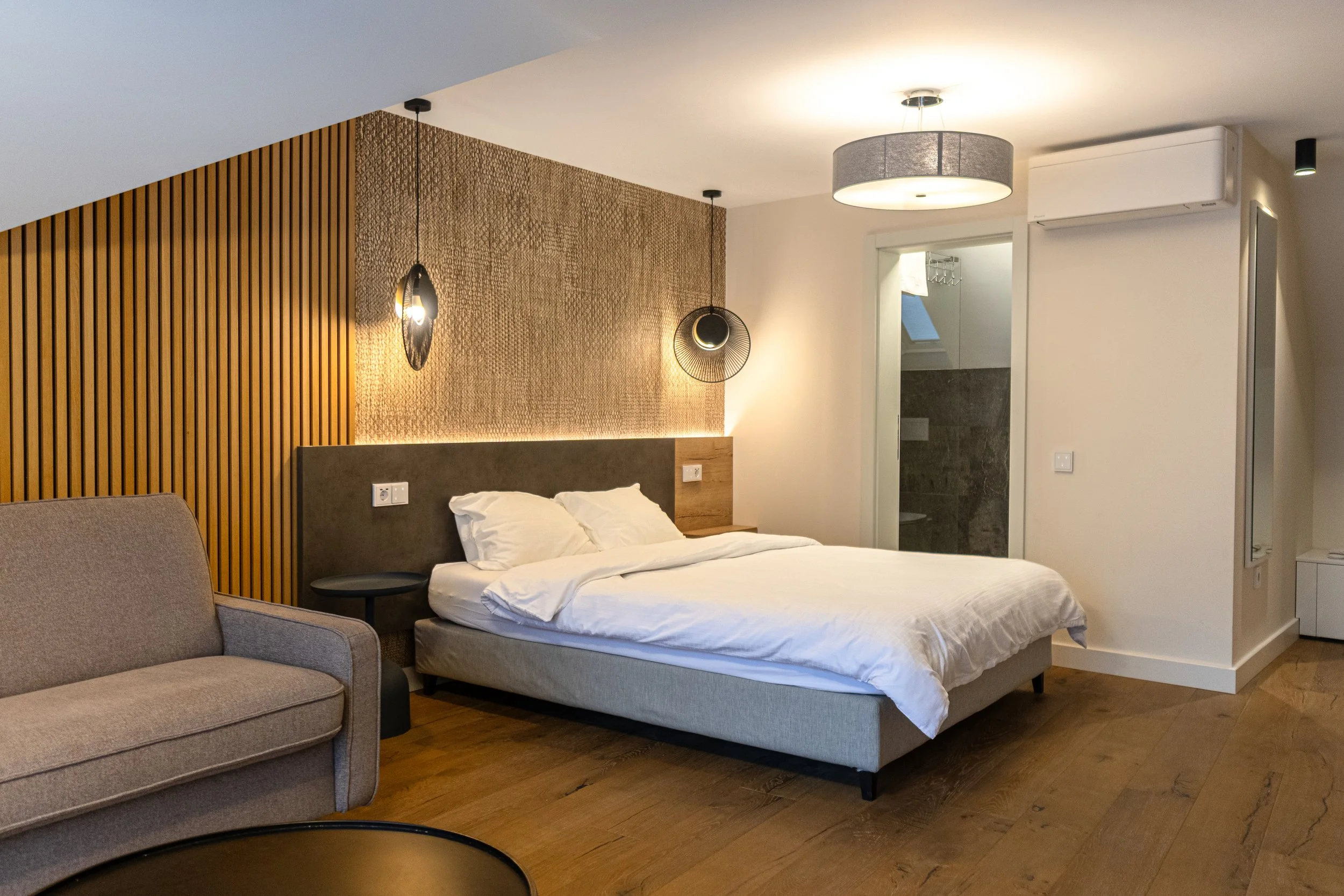 Modern hotel bedroom with a bed, a sofa, and stylish lighting fixtures, including hanging lamps and a ceiling light, hardwood floors, and a partial view of a bathroom.